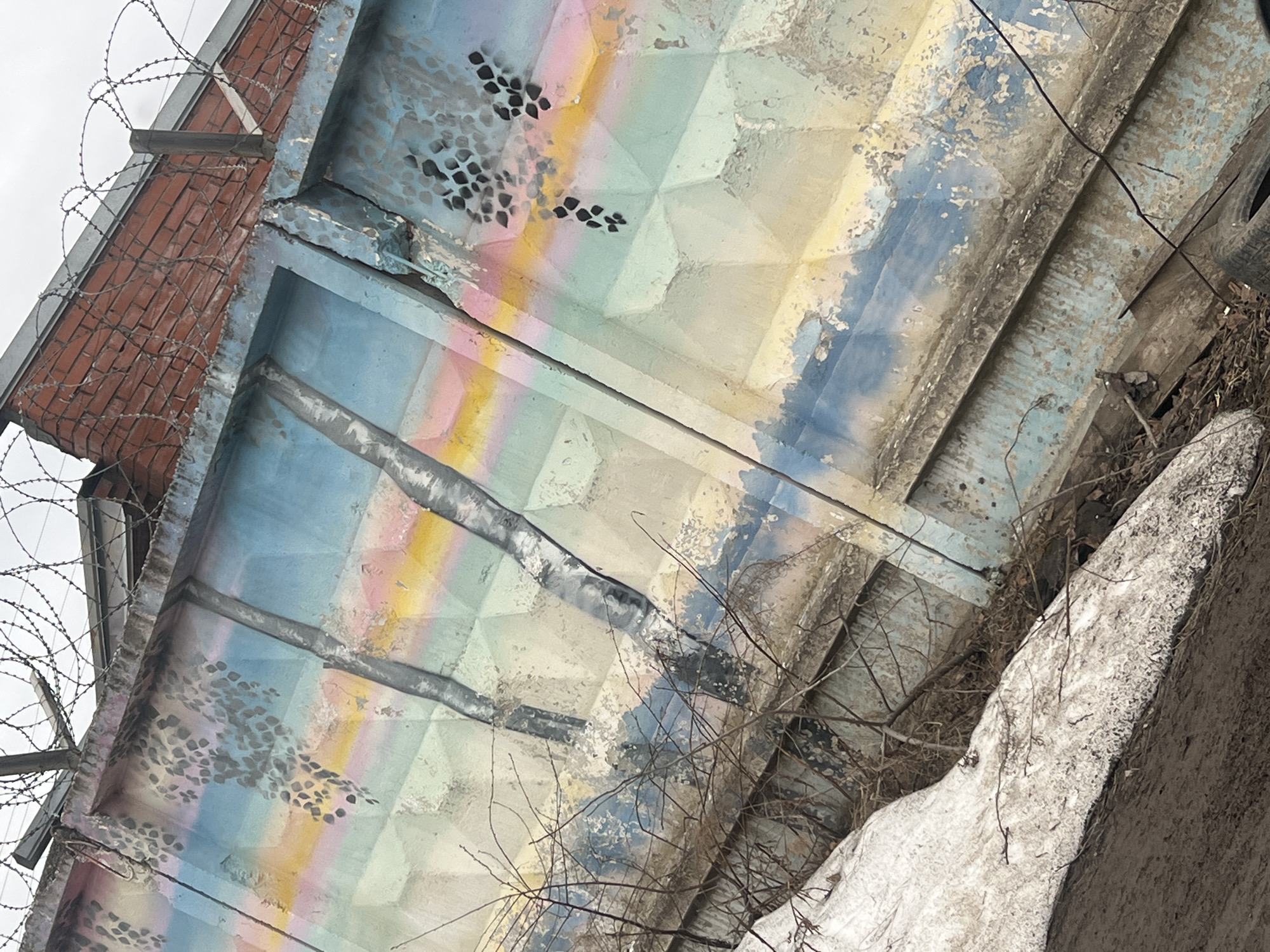 The image presents a close-up, upward-angled view of a weathered concrete structure, possibly a portion of a building or infrastructural element. The structure displays geometric paneling, painted with faded pastel colors including blue, yellow, and pink. There are areas of discoloration and decay, indicative of age and exposure to the elements. Black, abstract patterns are also present, appearing to be stenciled or painted on.
To the left, a brick building with barbed wire sits above the concrete structure, suggesting a secured perimeter. A tangle of bare branches and what appears to be a snow-covered rocky surface occupies the lower right portion of the frame. The scene is likely in an urban environment. Based on the prompt, this location is in the city of Chaikovsky, Russia. No people are visible in the frame.