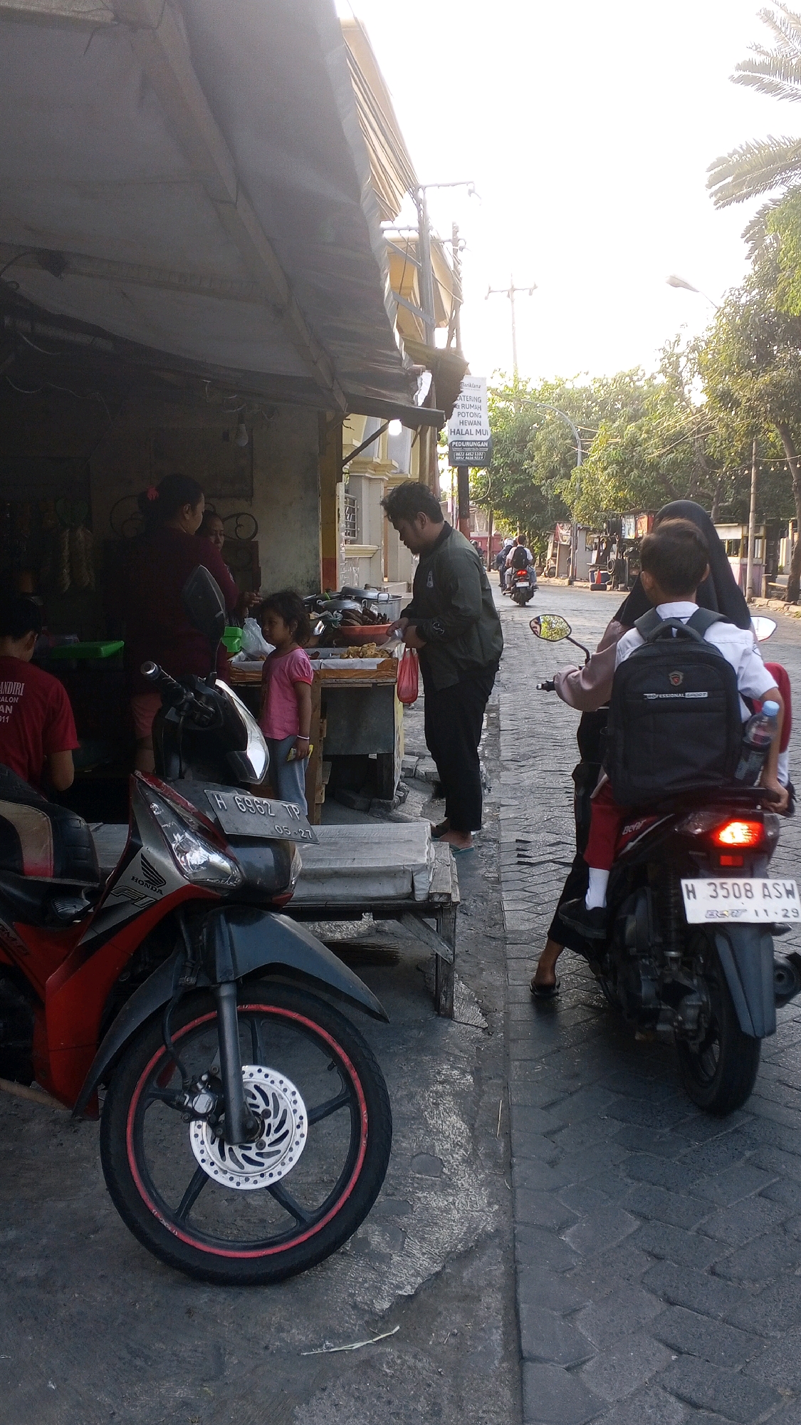 In a street scene in Semarang, Indonesia, a street vendor's stall is visible, with a canopy overhead. A red and black motorcycle is parked in the foreground. Several people are present: a man in a dark jacket is interacting with a young girl in a pink shirt, who is standing near a table displaying food items. Another woman is seen behind the girl, and a person in a red shirt is seated on a motorcycle on the left. In the background, a woman wearing a hijab is riding a motorcycle with a child wearing a backpack as a passenger. The street is paved with cobblestones, and trees line the side of the road. A sign in the background reads "CATERING RUMAH POTONG HEWAN HALAL MU".
