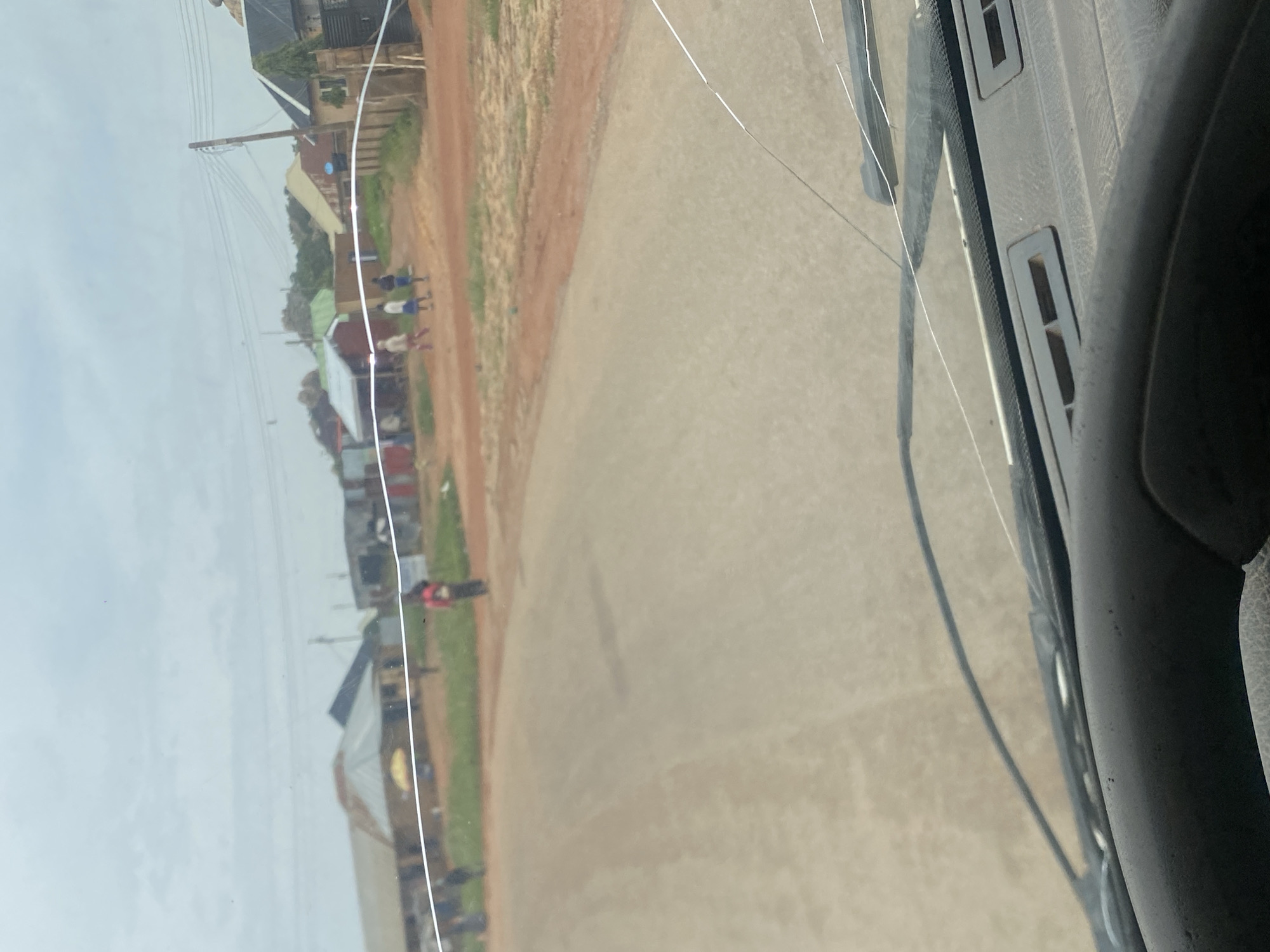The image depicts a dusty, unpaved road in what appears to be a rural or semi-urban area of DU, Nigeria. The sky is overcast, suggesting a cloudy day. Several small buildings with corrugated metal roofs line the right side of the road, some appearing to be shops or residences. In the background, there are hints of more structures and vegetation.
Several individuals are visible on or near the road. In the mid-ground, a person wearing a red top is standing on the right side of the road. Further to the left, a group of three younger individuals, likely children or teenagers, are walking away from the camera. One is wearing a light-colored top and blue shorts, another a red top and dark bottoms, and the third a dark top and blue shorts. On the far left, two individuals are partially visible, one seemingly wearing dark clothing.
The foreground of the image is dominated by the interior of a vehicle. A dark steering wheel is visible at the bottom, and a portion of the dashboard with air vents is present. The windshield of the vehicle is cracked, with visible lines distorting the view of the outside scene. A windshield wiper is also visible. The overall impression is a candid shot taken from inside a moving vehicle, capturing a slice of everyday life on a Nigerian road.