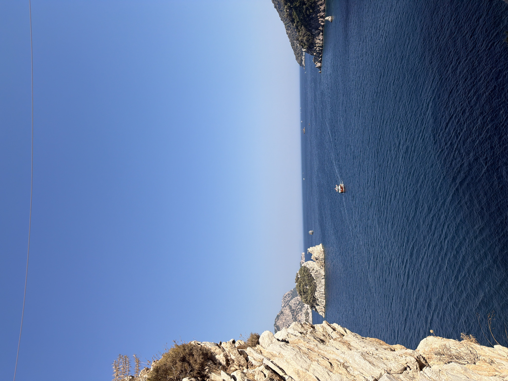 This is a high-angle, full shot of a blue sea with islands and boats, taken on a sunny day. In the foreground on the left, a large, textured, rocky cliff face dominates the left side of the frame. The cliff is lit by the sun, highlighting its rough surfaces and sparse vegetation.
Beyond the cliff, the deep blue sea stretches towards the horizon. Several small, rocky islands with green vegetation dot the water. The largest of these islands is located in the middle-ground, with a cluster of trees on its summit.
A few boats are visible on the water. One boat, a small white vessel with a red roof, is sailing in the middle of the frame. Further in the distance, other boats appear as small specks on the sea, suggesting a moderate level of activity.
The background shows a hazy coastline with green, tree-covered hills. The sky is a clear, cloudless blue, indicating a bright and clear day. A thin, dark wire stretches horizontally across the top of the frame, possibly a power line or part of a zip line.
The scene evokes a sense of a coastal resort or a scenic natural landscape, likely within the vicinity of Marmaris, Turkey, known for its beautiful coastline and islands.