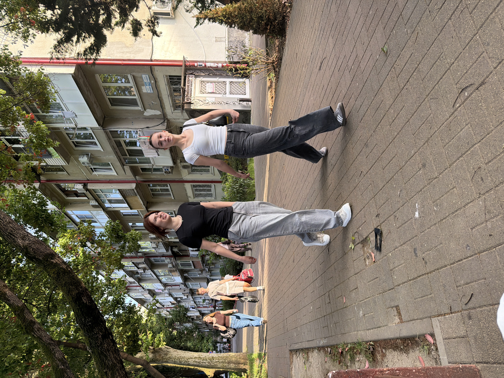 Two young women are captured in a full-body shot, posing dynamically on a brick-paved sidewalk. The woman in the foreground, wearing a black t-shirt and light gray jeans, leans back slightly with one leg extended. She has reddish-brown hair and is looking upwards. Behind her, another woman in a white t-shirt and dark gray jeans stands with her body angled, one leg forward. She has dark hair and is looking towards the camera. Both women are wearing sneakers.
The scene is an urban environment, likely in Sochi, Russia, as indicated by the architecture of the apartment building in the background. The building has multiple balconies and windows, suggesting a residential area. Lush green trees, characteristic of a warm climate, frame the left side of the image, their branches casting shadows. The lighting suggests it is daytime, possibly late afternoon, with the sun casting distinct shadows. The overall mood appears relaxed and casual, with the subjects appearing confident and playful in their poses. There are a few other people visible further down the sidewalk, and some bags are placed on the ground. No visible text is discernable in the image.