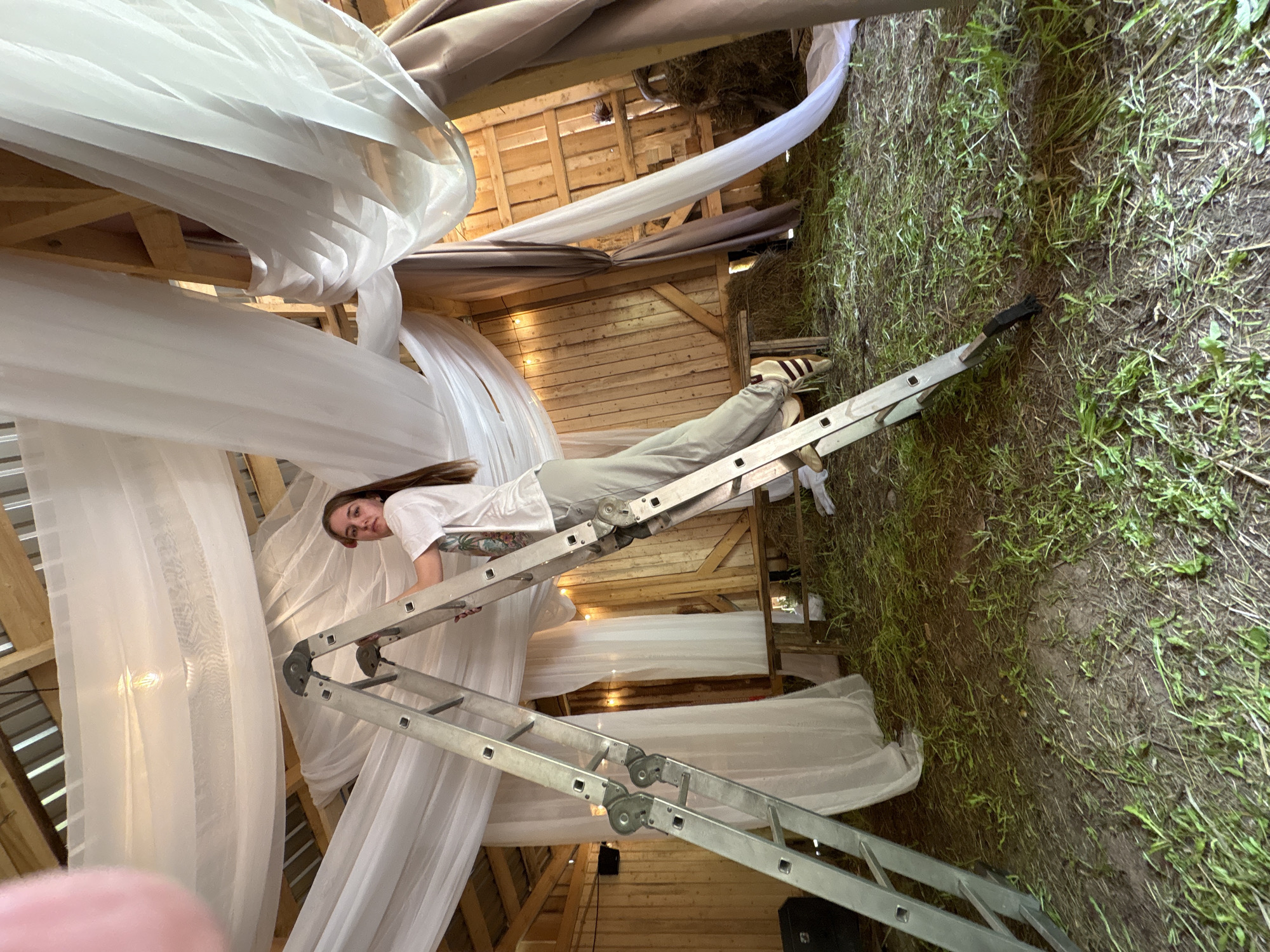 The image shows a woman standing on a metal ladder inside a wooden structure, possibly a barn or shed. The interior is decorated with white and beige fabric draping from the ceiling. The floor outside the structure is covered with grass and vegetation. The image was not taken in Sibir', Russia, as this information cannot be deduced from the image.