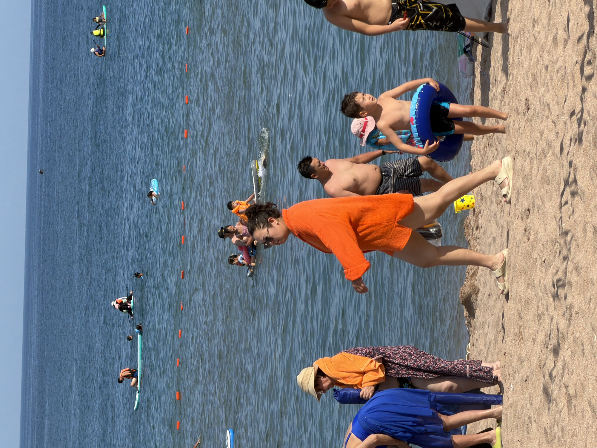 The image captures a sunny day at a beach with many people enjoying the water and sand. In the foreground, several individuals are walking on the beach. A woman in an orange cover-up walks away from the camera, while a child in swim trunks holds a blue inner tube. To the left, a person in a blue dress and another in an orange towel are partially visible.
In the background, the ocean is filled with activity. Several people are on paddleboards, including a man wearing an orange life vest. Other individuals are swimming or wading in the water. A marked swimming area is delineated by orange buoys. The sky is clear and blue, indicating fair weather. The overall scene depicts a typical summer day at a crowded beach resort.