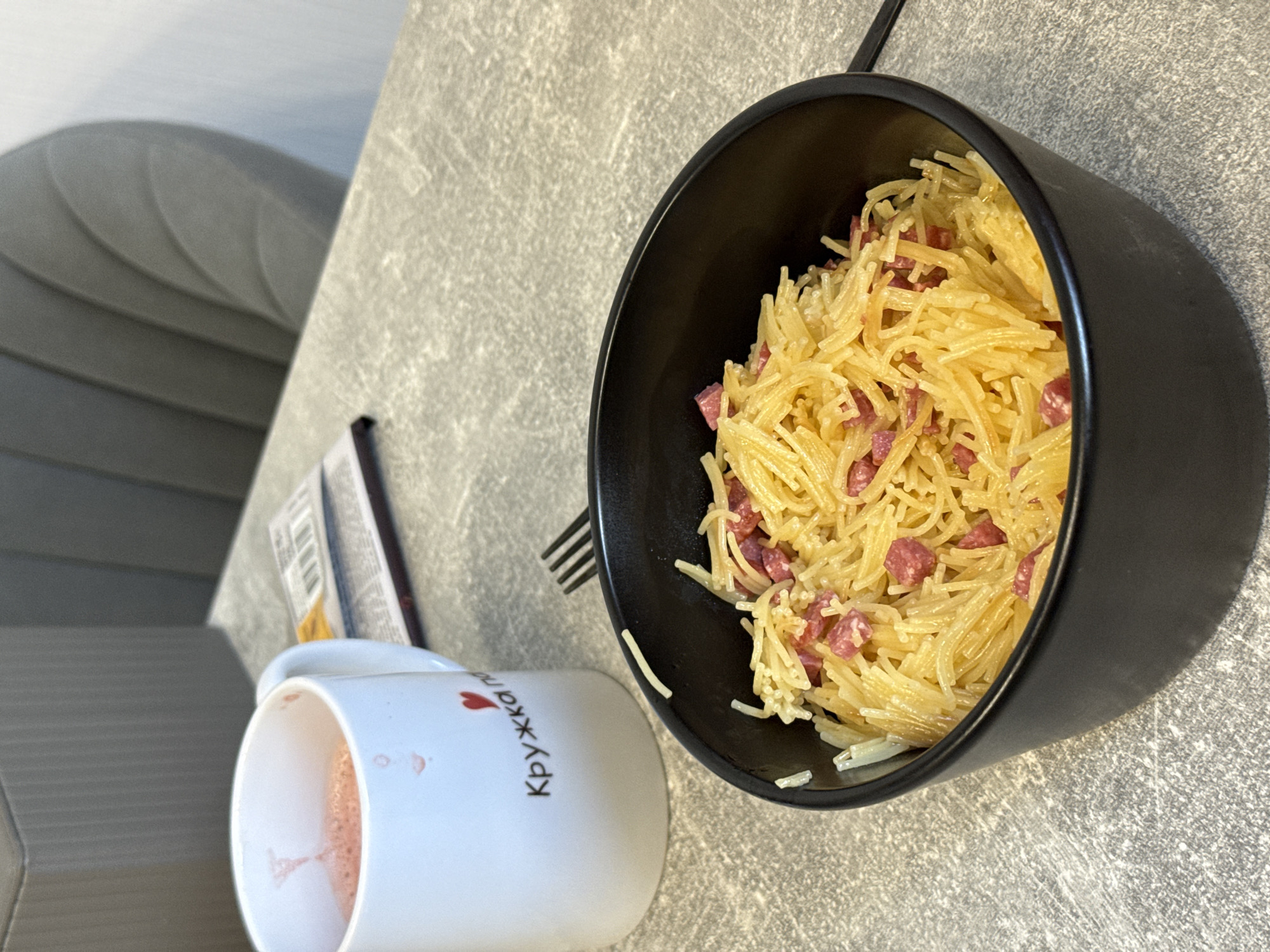A close-up, eye-level shot shows a bowl of spaghetti with diced meat and a white mug containing a pink beverage. The food is served in a black bowl, positioned in the foreground. The mug, to the left of the bowl, has red writing and a small heart design. A fork is visible to the right of the bowl. The objects are placed on a grey, textured table, which appears to be in a dining area or kitchen. In the background, a muted grey chair and a portion of a grey structure are visible. The scene is evenly lit, with no strong shadows. The image does not provide any specific indicators of its location in Lyubertsy, Russia.