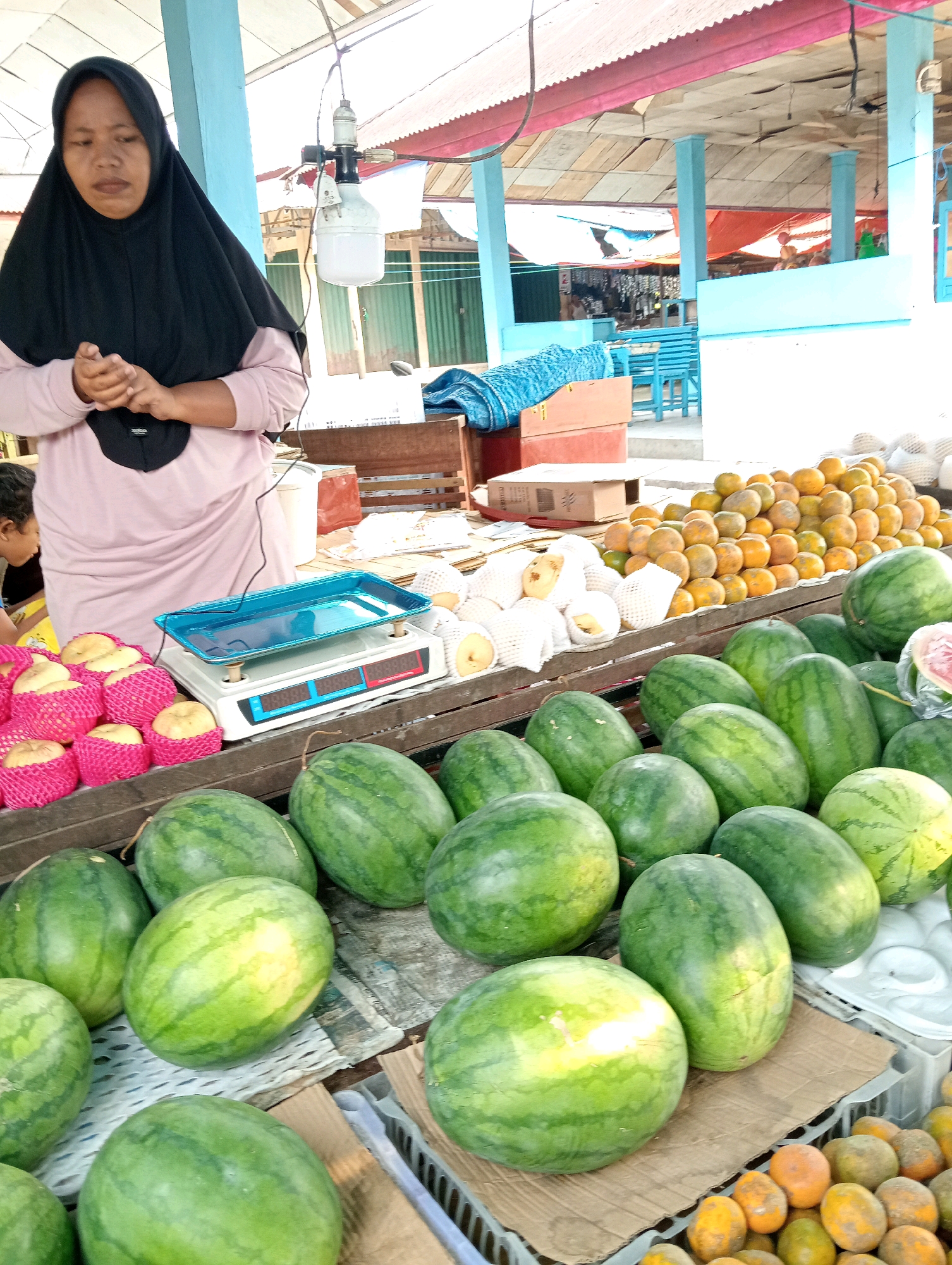 In Pariaman, Indonesia, a woman wearing a black hijab and a light pink long-sleeved shirt stands at a fruit stall. Her hands are clasped in front of her. In front of her is a blue-tray scale on a white electronic scale. Piles of apples, wrapped in pink foam netting, are to the left of the scale. Behind the apples and scale are stacks of oranges. In the foreground, numerous watermelons are displayed on wooden crates. Other fruits, possibly pears, wrapped in white foam netting, are visible on a crate behind the oranges. The stall is covered by a corrugated metal roof with pink trim, supported by blue columns. In the background, there are glimpses of other market stalls and structures.