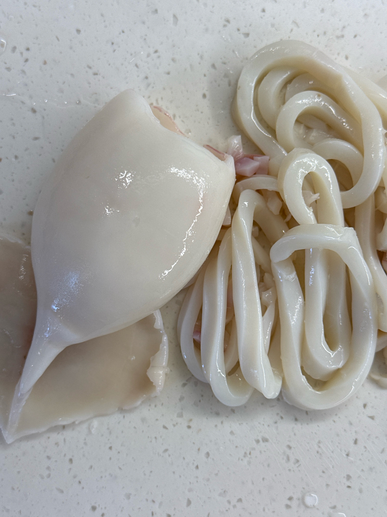 The image displays a close-up, overhead view of raw, prepared squid on a speckled white countertop. To the left is a whole squid mantle, appearing plump and smooth, with a tapered end. To the right, the squid has been sliced into rings, which are piled together. The squid is a pale, translucent white color with a slightly pinkish hue visible in some areas, particularly around the edges of the rings. The countertop has a subtle, uniform texture with small, dark speckles. There is a slight sheen of moisture on both the squid and the countertop, suggesting it has recently been cleaned or rinsed. The lighting is bright and even, creating minimal shadows. The image is taken in a static, indoor environment, likely a kitchen. There is no information within the image to indicate a specific location within the city of Lyubertsy, Russia.