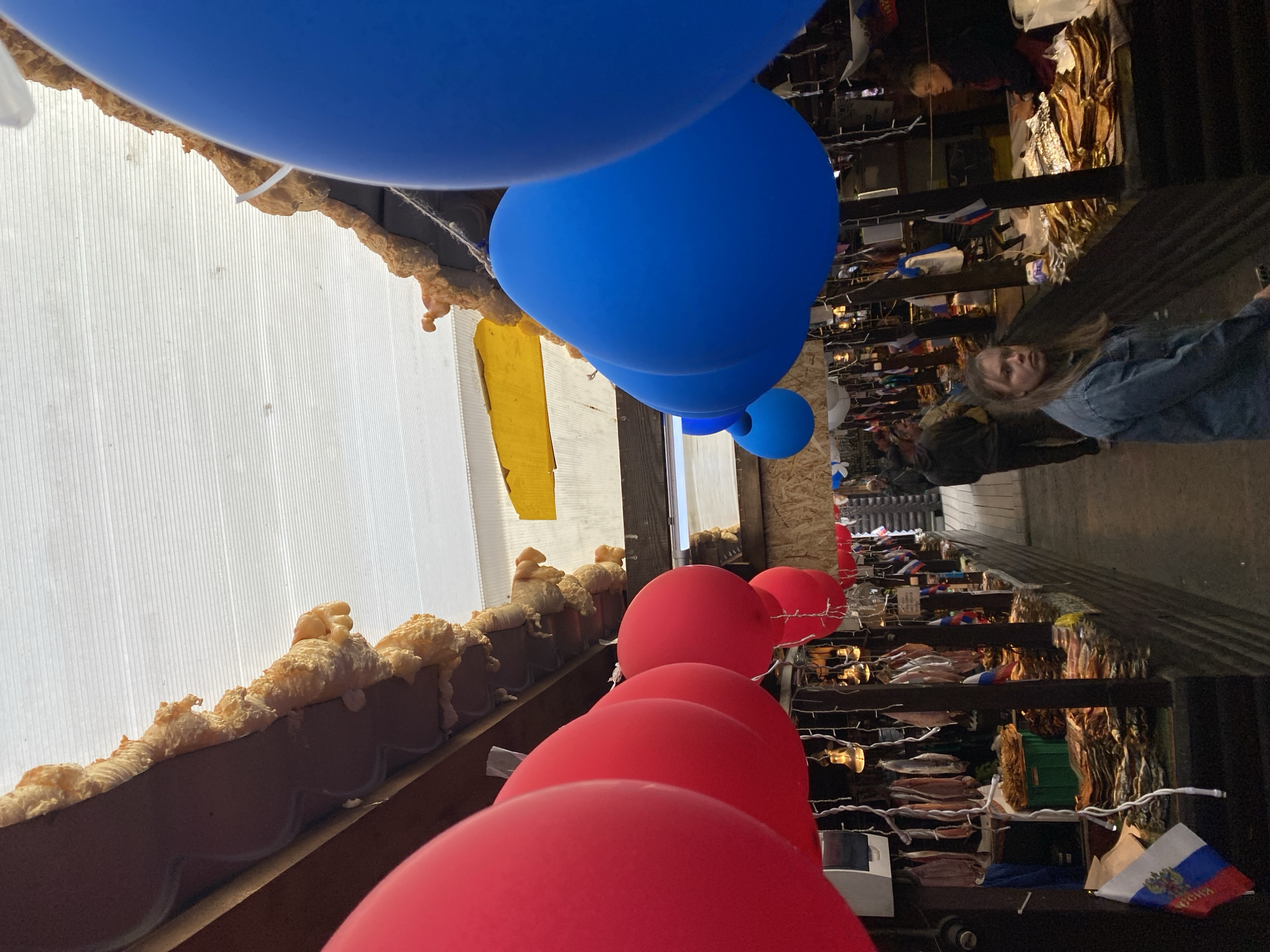 The image shows an outdoor market stall in Listvyanka, Russia. Red and blue balloons are suspended from the ceiling of the stall, which is covered with a white translucent material. The stall is filled with various goods, including dried fish, which are piled high on shelves and tables. Several people are browsing the stalls, including a woman in a denim jacket in the foreground and a vendor in the background. Russian flags are visible throughout the market. The overall impression is one of a bustling marketplace, possibly during a festival or celebration.