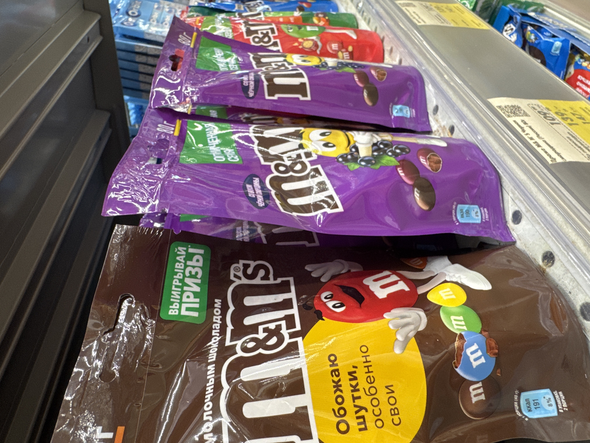 This media file displays a close-up, slightly angled shot of several bags of M&M's candy arranged on a store shelf. The dominant colors are purple, brown, and yellow from the candy packaging, with some blue and green also visible. The products appear to be M&M's candies, with some bags featuring purple packaging and depictions of M&M characters and blueberries, suggesting a "blackcurrant" flavor. Another bag in the foreground is brown and features the M&M's logo and characters, along with Russian text.
Visible text on the packaging includes "ВЫИГРЫВАЙ ПРИЗЫ" (Win Prizes), "молочным шоколадом" (with milk chocolate), and nutritional information like "ккал 191" (kcal 191) and "8%". Other text visible on the purple bags includes "80г" (80g) and descriptions possibly related to the flavor. There are also price tags visible on a shelf above the candy, showing prices like "139" and "147".
The setting is clearly a retail environment, likely a supermarket or convenience store, indicated by the shelves and the organized display of products. The lighting is bright, typical of indoor retail lighting, and there are no discernible environmental cues related to time of day or weather. The focus is entirely on the candy products, with no people or animals present. The overall impression is one of commercial display, highlighting the product's branding and potential promotions.