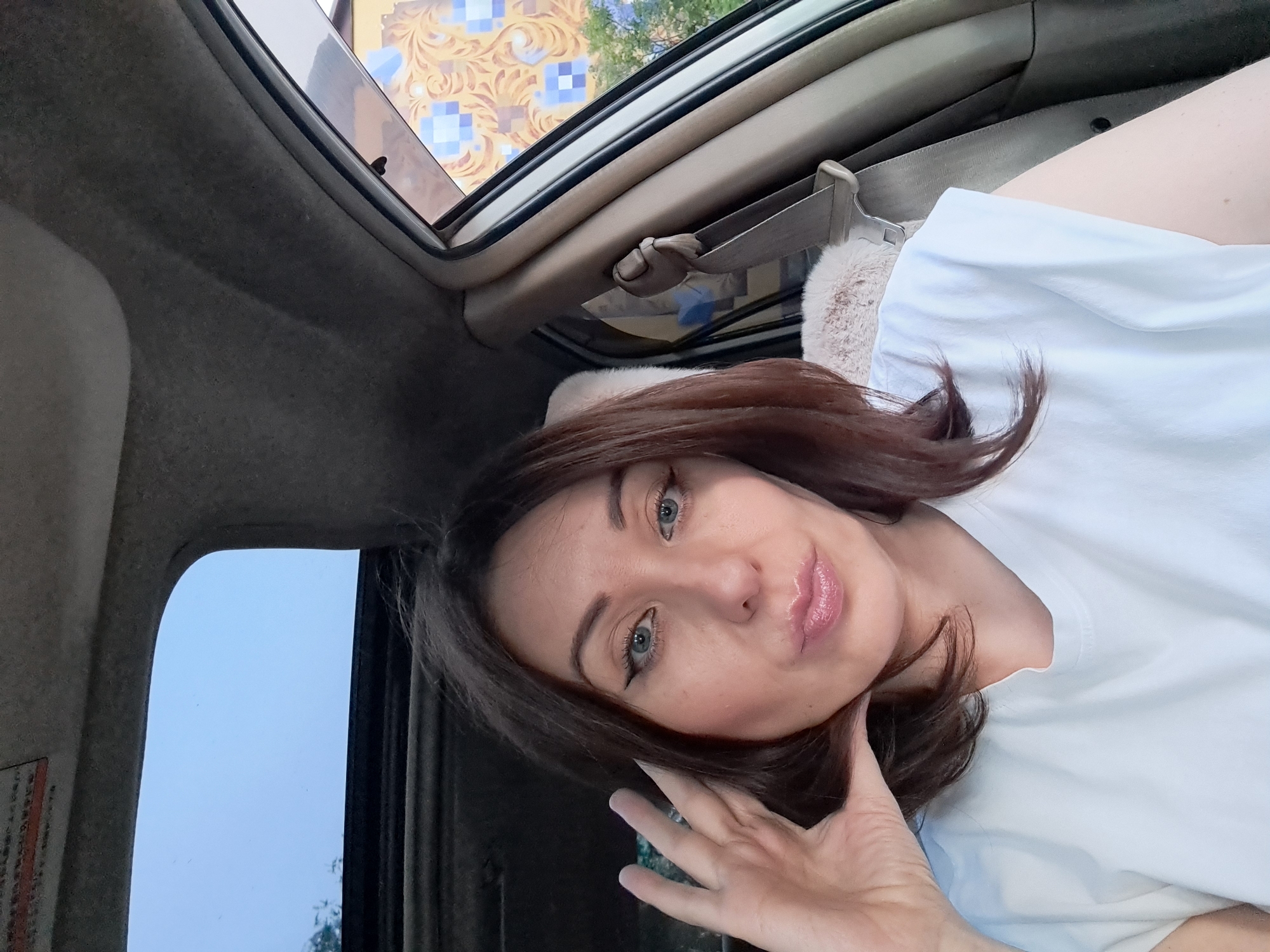 A woman is sitting in the passenger seat of a car, with a sunroof open above her. She has brown hair and is wearing a white t-shirt. She is looking at the camera and has her left hand raised to her face. The interior of the car is visible, including a seatbelt and part of the dashboard. Outside the car window, a building with a patterned facade is visible. The location is Irkutsk, Russia.