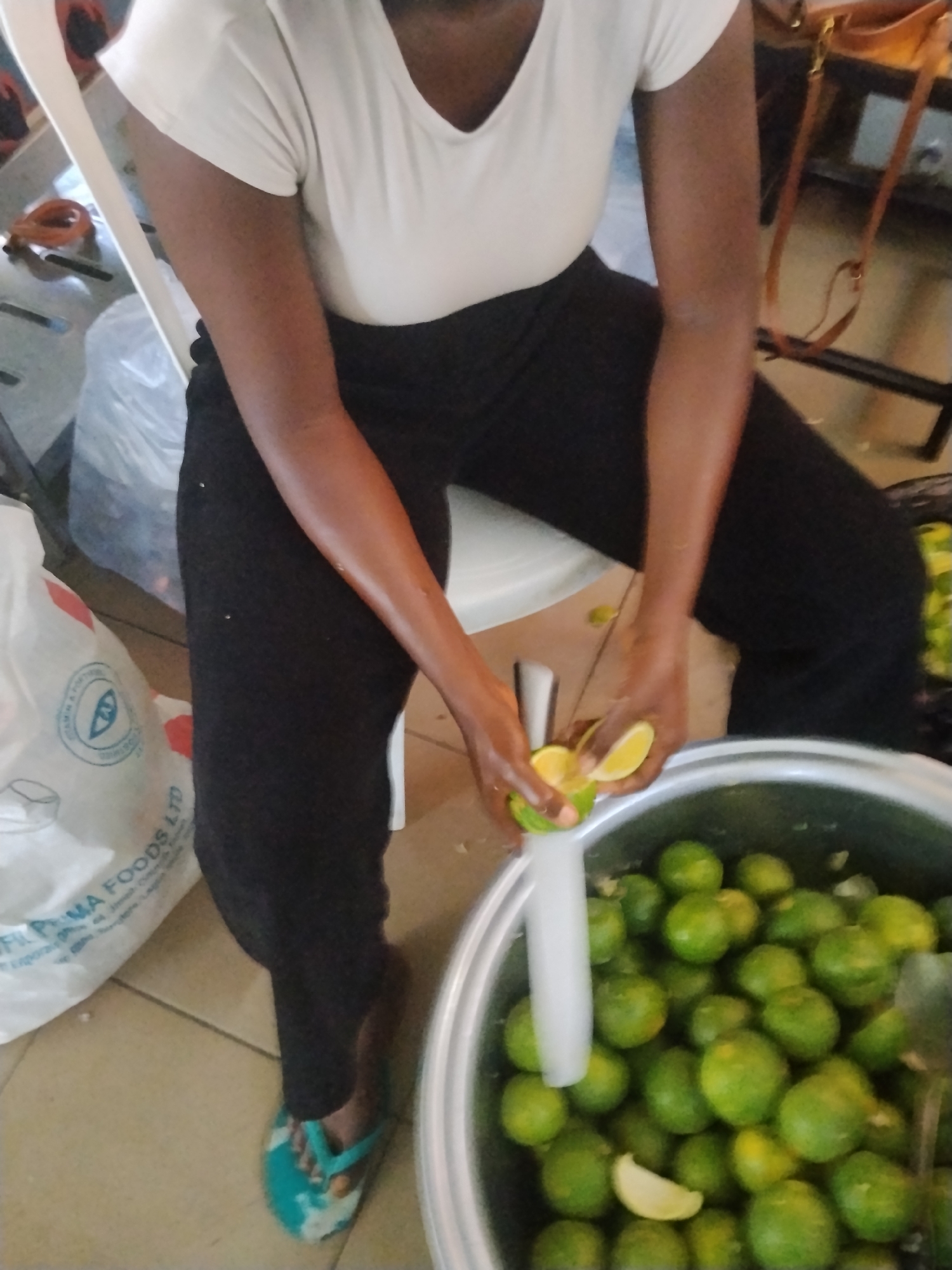A person wearing a white t-shirt and black pants is sitting on a white chair and preparing limes. They are holding a lime in their hands, squeezing it over a large silver bucket filled with more limes. The scene appears to be indoors, possibly in a kitchen or market stall, with a plastic bag and other objects visible in the background. The floor is tiled. This scene is located in the city of Howayi, Nigeria.