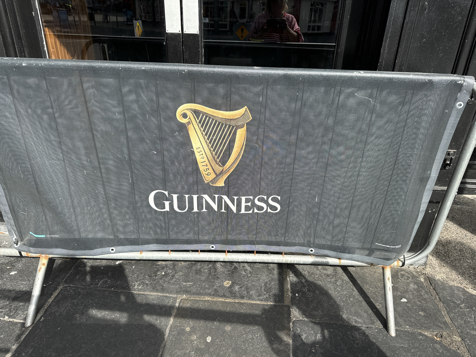 A black mesh barrier displays the Guinness logo and text. The logo, a golden harp, is positioned above the word "GUINNESS" in white, sans-serif font. The barrier is supported by rusty metal poles and is placed on a stone pavement outside a building. A reflection in the building's glass window shows a person wearing a striped shirt, holding a phone. This scene is located on Georges Street, Waterford, Ireland.