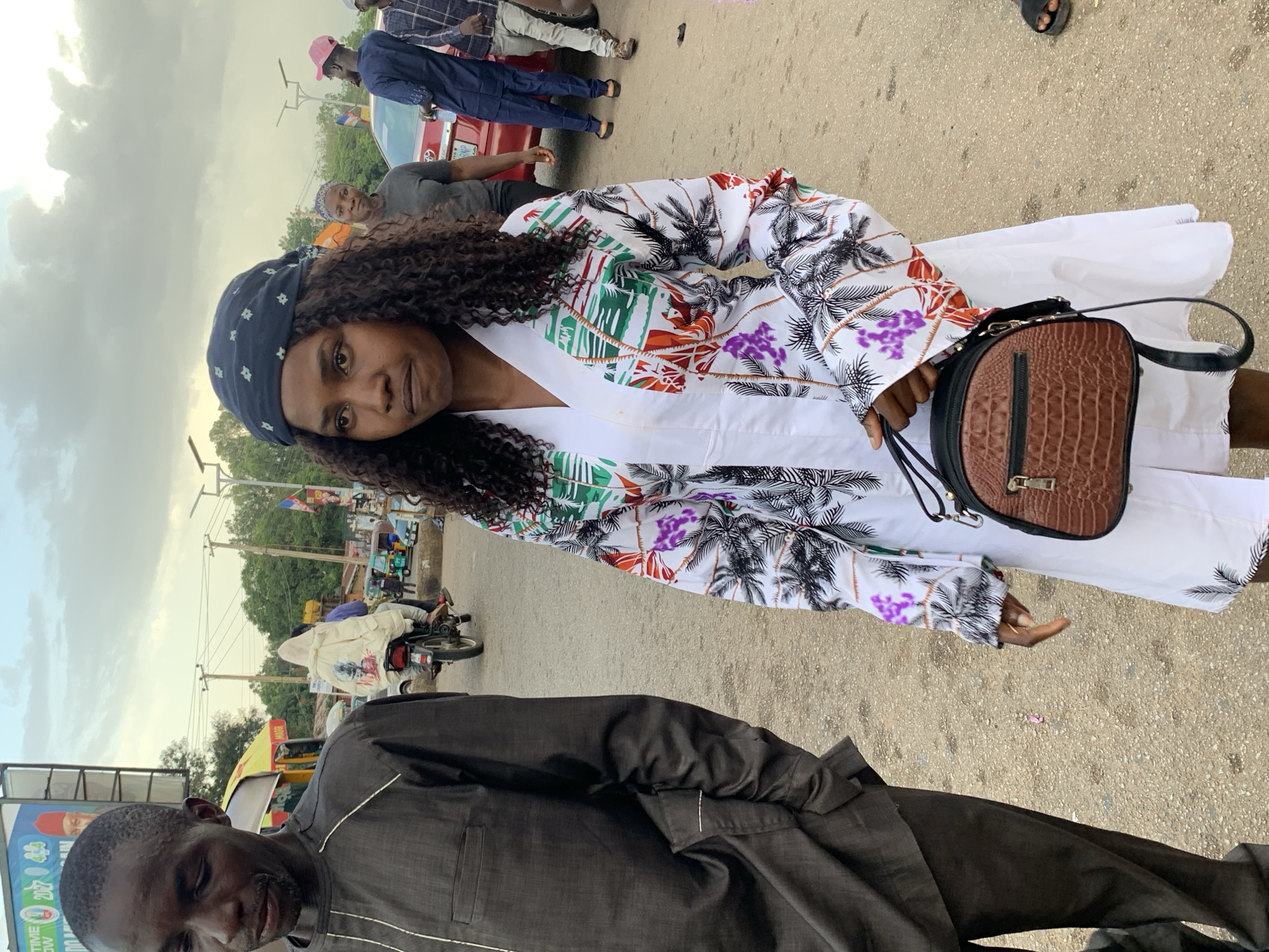 The image depicts a street scene in Jos, Nigeria, likely during daytime with overcast skies. In the foreground, a young woman with dark, curly hair, wearing a headscarf and a white dress with a colorful tropical print on the sleeves, holds a brown, crocodile-textured handbag. To her left, partially in frame, is a man wearing a dark grey traditional outfit. In the background, other individuals are visible, including a person on a motorcycle, and a red car. The setting appears to be a moderately busy street with a paved surface, bordered by vegetation and some roadside structures. The overall atmosphere is indicative of a typical urban environment in Nigeria.