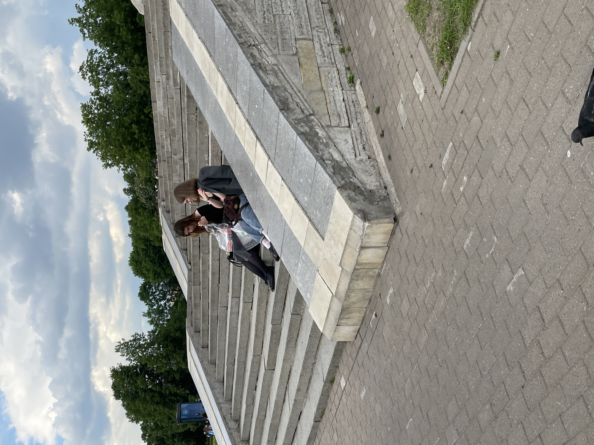 The image captures a daytime scene in what appears to be a public park or urban area in Saint Petersburg, Russia, centered around a wide set of stone stairs. The photograph is oriented vertically, showing the stairs leading diagonally upwards from the lower left to the upper right.
Two young women are visible on the stairs, positioned roughly in the middle of the frame. One woman, with long brown hair, wears a dark top and blue jeans, carrying a dark shoulder bag. The other, partially obscured, appears to have lighter hair and a lighter-colored jacket or top. Both are leaning forward, engrossed in looking down at something small held by one of them, likely a smartphone, suggesting they are resting or taking a break.
The stairs are made of grey stone or concrete, featuring a distinct lighter-colored strip running horizontally along the balustrade. To the right of the stairs, a pathway is paved with grey interlocking bricks laid in a herringbone pattern. Small patches of green grass are visible between some of the pavers and along the edge where the path meets another surface. A pigeon's head and partial body are visible on the far right edge of this paved area, looking towards the left.
In the background and extending above the stairs, numerous lush green trees create a natural canopy. The sky above is bright but partly cloudy, with several white and grey clouds against a light blue backdrop. The overall impression is one of a casual, relaxed moment in a serene public space. No discernible text is visible on any objects, signs, or clothing.