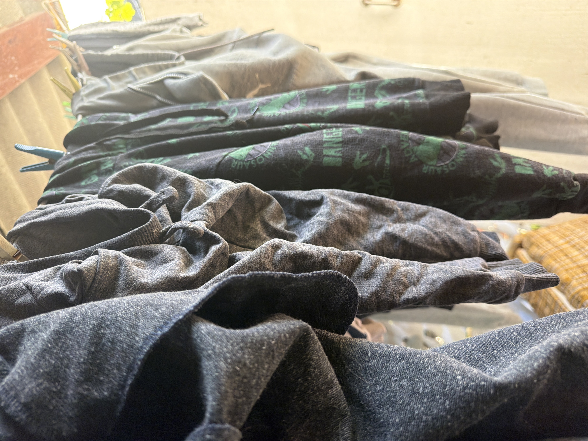 The image shows a stack of folded clothing. The bottom item is a dark gray sweatshirt or sweater. Above it is a lighter gray sweatshirt or sweater. On top of that is a black shirt or sweatshirt with green dinosaur print. A light gray garment sits on top, and another item is barely visible above. The items appear to be stacked indoors and may be freshly laundered, given the visible clothespin. No location or people are discernible in the image. The image does not contain information that relates to Agoy, Russia.