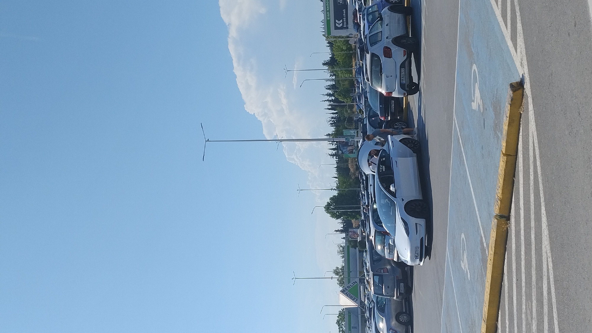 A wide-angle, full-shot photograph captures a crowded outdoor parking lot on a clear, sunny day in Pylaia Municipal Unit, Greece. The foreground shows a section of the asphalt parking lot marked with blue and white lines, including a blue-painted space designated for disabled parking with a white wheelchair symbol. A low, yellow curb runs along the edge of the parking bay.
In the middle ground, numerous cars are parked in rows. A prominent white Tesla Model 3 is parked in the foreground, facing left, with its rear trunk open. Three individuals are visible near the Tesla; two men and one woman. One man, wearing a grey t-shirt and shorts, stands beside the open trunk. Another man, wearing a white t-shirt, is partially visible behind the Tesla. A woman, wearing a white top and dark shorts, is also near the open trunk. Other vehicles in the middle ground include a white Dacia Sandero, a silver hatchback, and a dark-colored SUV.
In the background, several tall streetlights are visible. Beyond the parking lot, a building with a green and white sign that reads "LEROY MERLIN" is present. Further in the distance, a line of trees and a slightly hilly landscape are visible under a bright blue sky with scattered white clouds. The overall scene suggests a busy commercial area, likely a shopping center or retail park.