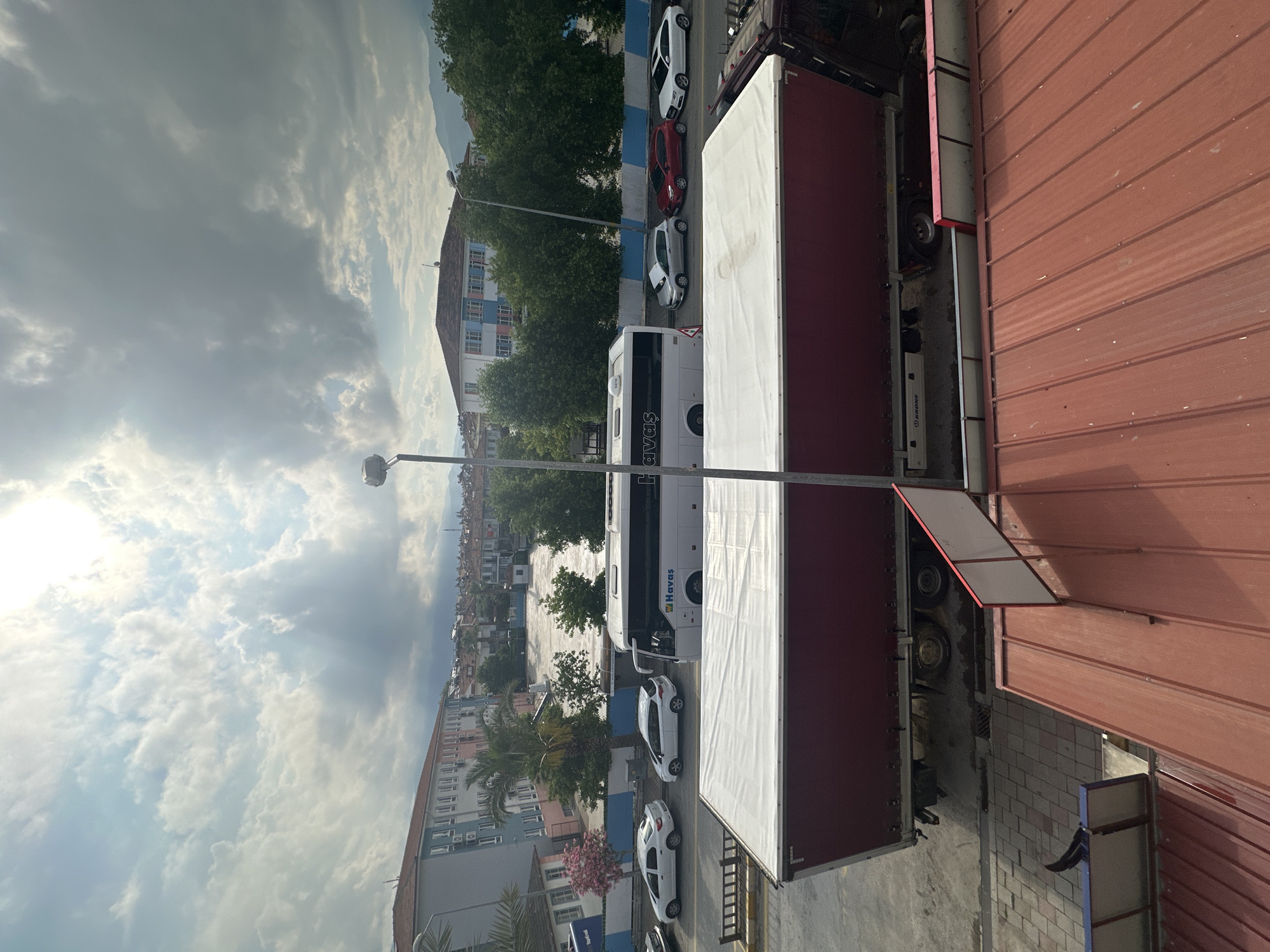 This image shows a street scene in Fethiye, Turkey, viewed from an elevated position, potentially a balcony or higher floor of a building. A Havaş bus is parked along the street, along with several cars. A large truck with a red cargo area is also visible, parked to the right. There are several multi-story buildings along the street, and trees are scattered throughout the area. The sky is cloudy with patches of sunlight. A streetlight stands tall.