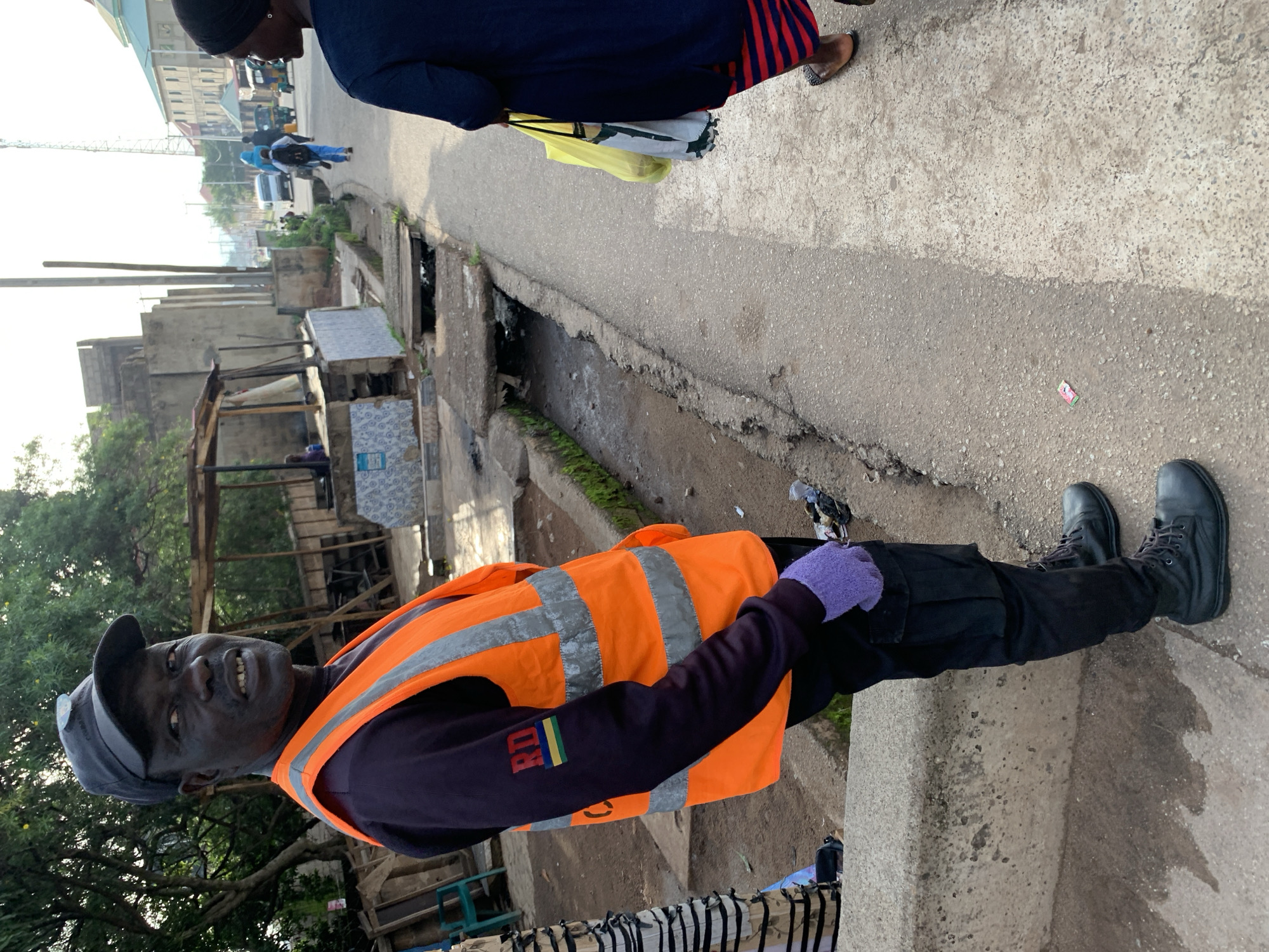 The image captures a street scene in Jos, Nigeria. In the foreground, a Black man wearing a dark blue long-sleeved shirt, black cargo pants, and black boots stands facing the camera. He wears a bright orange safety vest with reflective stripes over his shirt. On his left sleeve, there is a patch with the letters "RD" and horizontal stripes in green, yellow, and blue. He also wears a black baseball cap and light purple gloves.
To the right of the man, a Black woman is seen from the back, walking away from the camera. She wears a dark blue top and a red and black striped skirt. She carries a yellow plastic bag and a white and green patterned bag.
The environment appears to be an urban street with a paved sidewalk and a concrete drainage channel running alongside it. Buildings are visible in the background, along with some trees. Traffic, including a white van and several tricycles, can be seen further down the street, suggesting it is a public thoroughfare. The lighting indicates it is daytime, likely morning or afternoon.