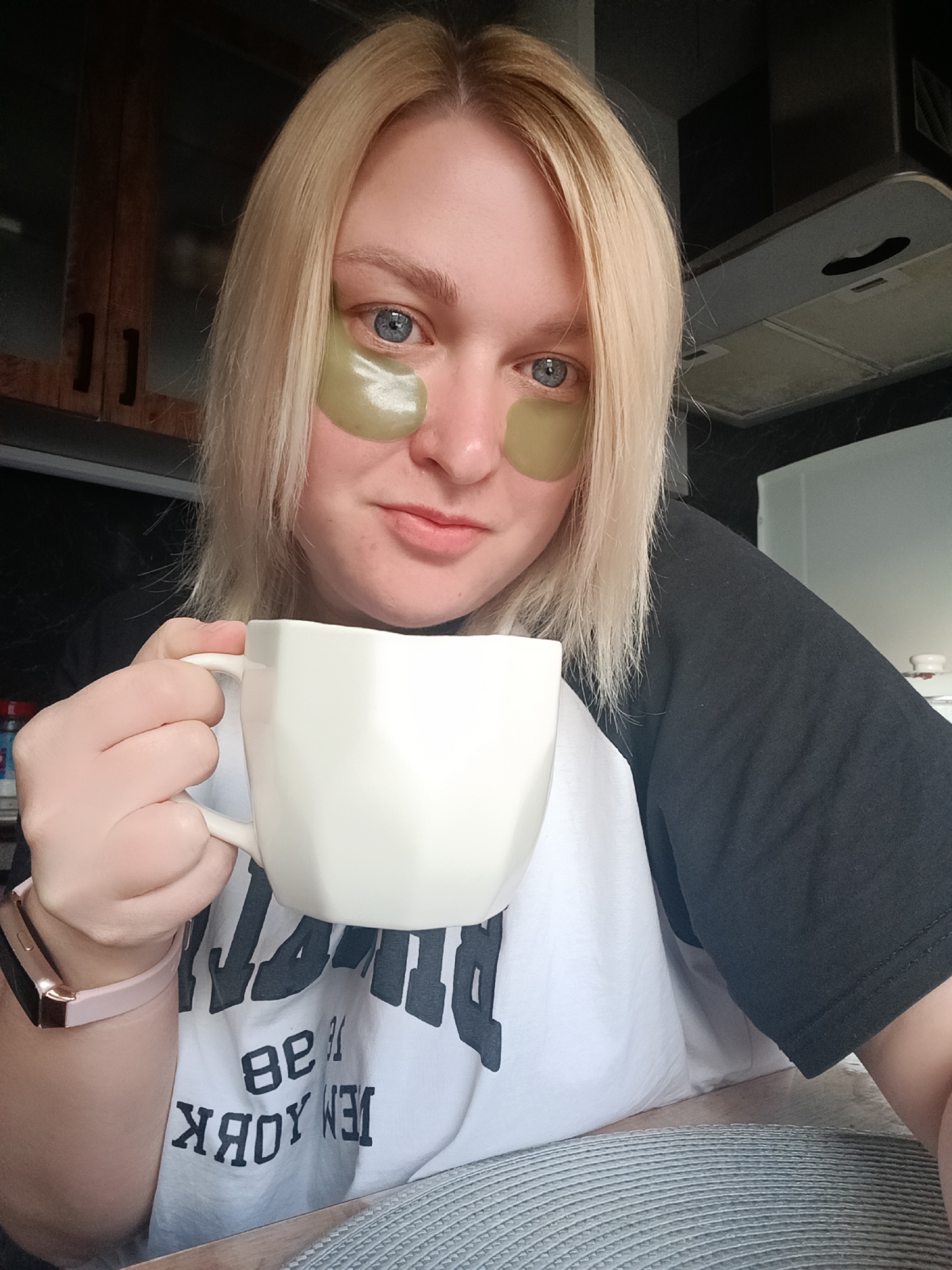 An indoor, well-lit selfie captures a person with blonde hair and blue eyes in what appears to be a kitchen or dining area, located in Chaikovsky, Russia. The individual, likely a woman, is wearing green-tinted hydrogel under-eye patches, indicating a morning skincare routine. She holds a faceted white mug in her right hand, suggesting she is drinking a beverage. On her left wrist, a pink smartwatch is visible. She looks directly at the camera with a neutral expression, hinting at a slight, soft smile.
Her attire is a black and white raglan-style t-shirt. Visible text on the white portion of the shirt, appearing upside down and reversed, clearly spells "NEW YORK". The background includes dark wooden cabinets on the left and a silver kitchen range hood on the right. A textured grey placemat is seen in the foreground. The overall impression is a relaxed, everyday moment during daytime.