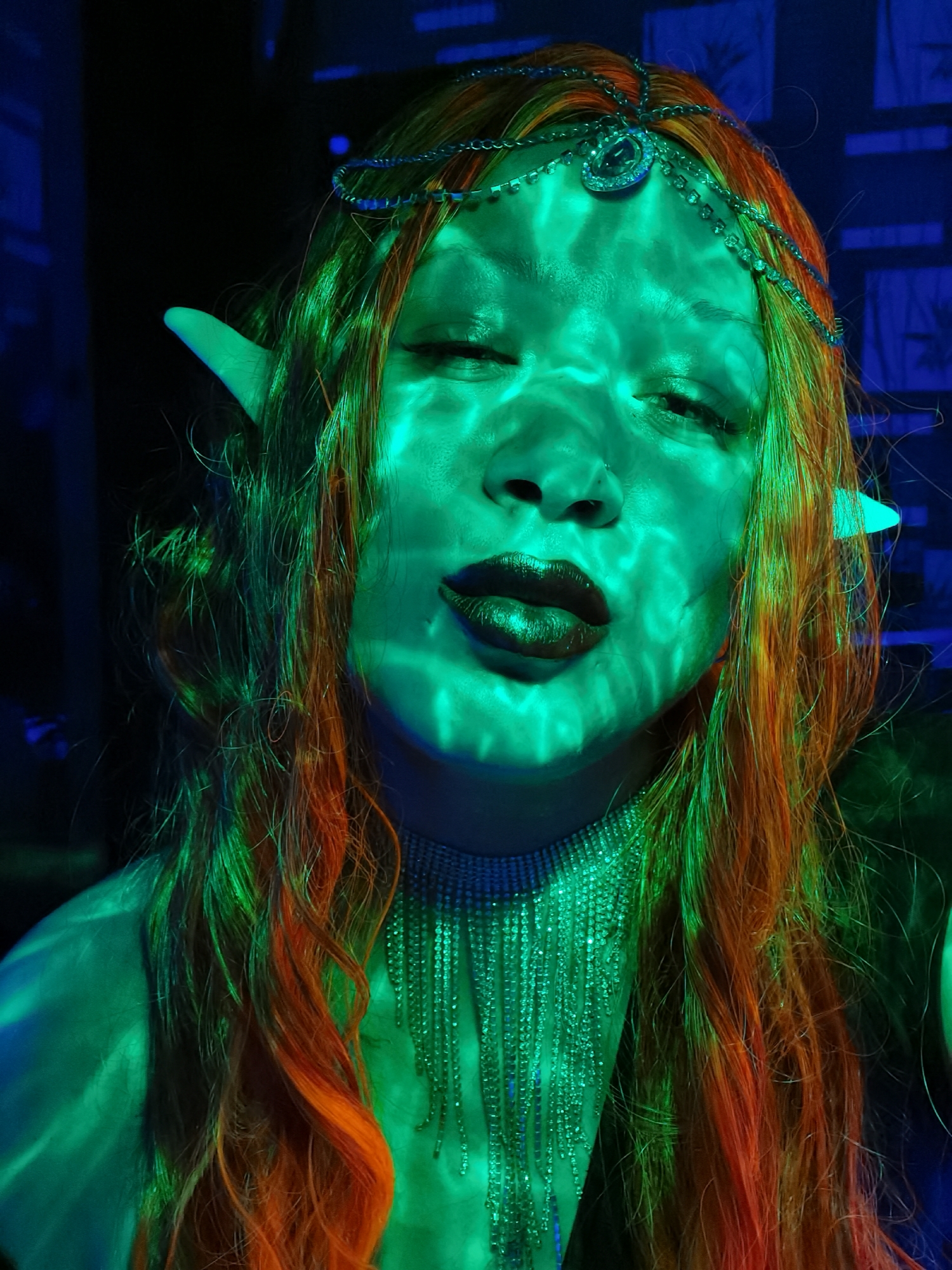 A close-up portrait shows a person with green skin, pointed ears, and closed eyes, posing with puckered lips. The person wears a rhinestone headpiece and a matching choker necklace with dangling strands. Their hair is long, flowing, and colored in shades of orange and green. Green light illuminates the face, casting patterned shadows. The background contains dark areas and hints of blue light, suggesting an indoor setting. The location in Chaikovsky, Russia, is not discernible from the image.