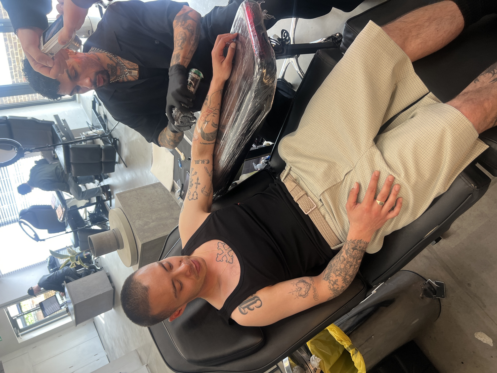 This media file captures a tattoo session in a modern, well-lit studio, likely located in London, United Kingdom. The primary subject is a man with short dark hair, lying on a black tattoo chair. He wears a black tank top and light-colored, checkered shorts with a beige belt. He has numerous tattoos visible on his arms, chest (a cross design), and legs. Text "ELIJAH" is visible on his right forearm and "C.H.M." is on his left forearm, along with other designs. He appears relaxed, with his eyes closed, while a tattoo artist works on his right arm.
The tattoo artist, a man with dark curly hair, a beard, and extensive tattoos on his arms and neck, is dressed in a black shirt, a silver chain, and black gloves. He is intensely focused on the tattooing process, using a tattoo machine and holding a green bottle, partially labeled "NITTRO," likely a tattoo supply. The client's arm rests on a black armrest wrapped in clear protective plastic.
In the upper left quadrant, a hand is visible holding a white digital camera, documenting the artist at work. The studio environment features large windows, suggesting daytime, and a clean, industrial aesthetic with concrete-like stands, modern furniture, and several other tattoo stations visible in the background. A yellow trash bag is also partially visible on the floor near the client's feet. The client wears a gold ring on his right ring finger.
