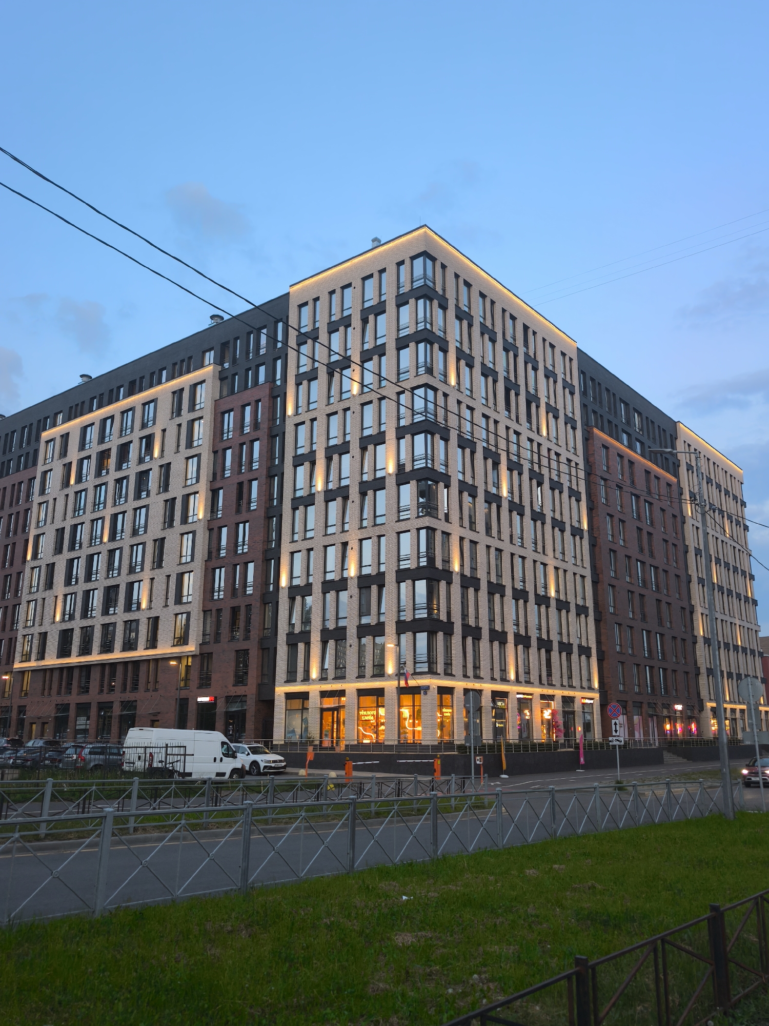 This is a full-frame, eye-level, outdoor shot of a modern, multi-story building in the city of Saint Petersburg, Russia, taken during twilight. The building features a facade constructed from a combination of light beige and dark reddish-brown bricks, with large, dark-framed windows. Accent lighting is installed along the edges and between floors, illuminating the facade. The sky above is a clear, pale blue with a few wispy clouds.
In front of the building, there is a street with a metal fence and a grassy median separating it from a sidewalk. Several vehicles are visible on the street, including a white delivery van, a white car, and other parked cars. Some businesses with illuminated storefronts are located at the ground level of the building. Power lines are visible crisscrossing the upper portion of the frame.