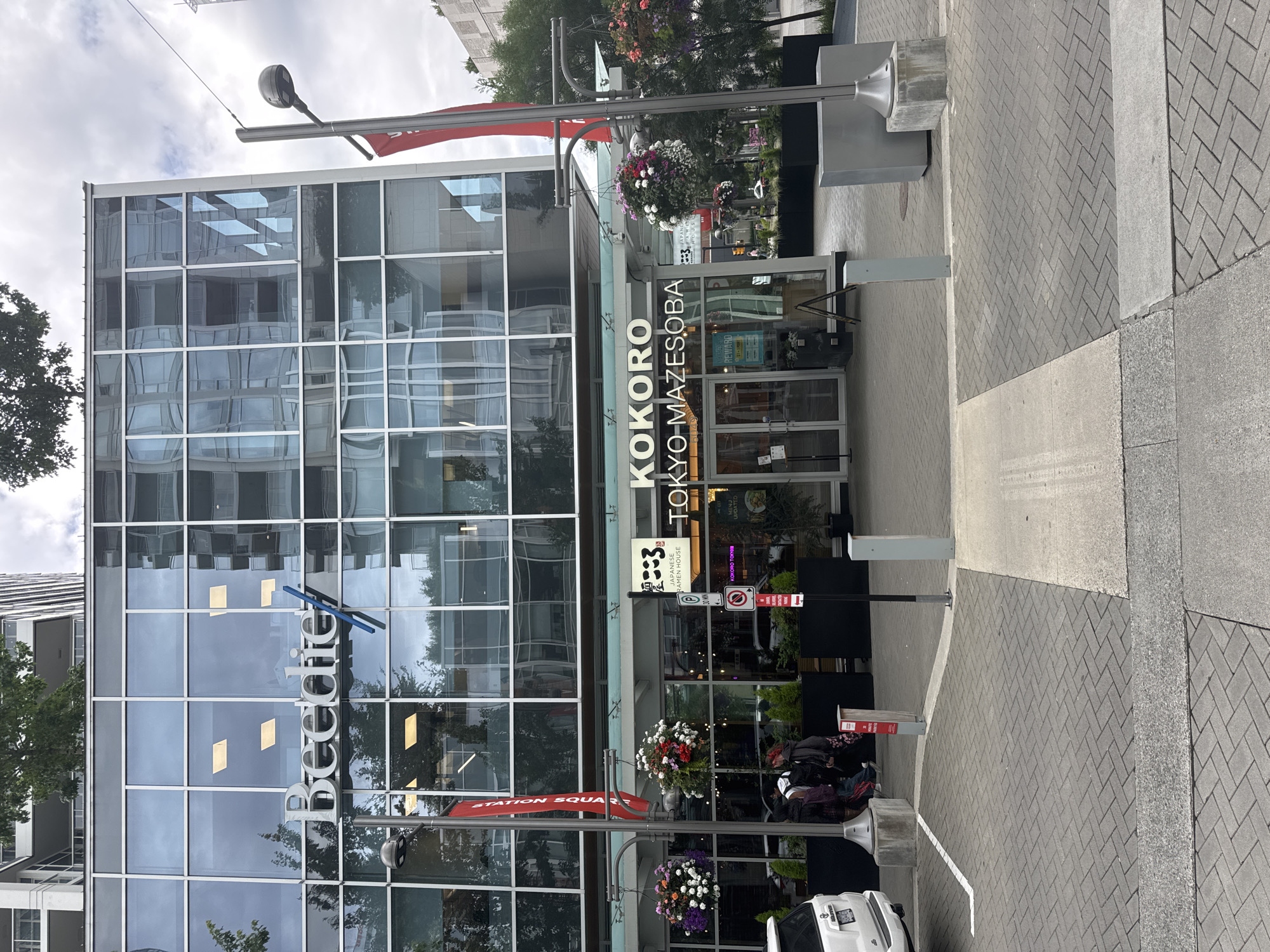 The image shows the exterior of a modern commercial building with a glass facade, featuring the "Beedie" logo prominently displayed. Below the glass facade, a restaurant with the name "KOKORO TOKYO-MAZESOBA" is visible. Red banners hang from lampposts, one of which reads "STATION SQUARE." Several potted plants and hanging flower baskets are placed outside the restaurant. In the foreground, there is a paved walkway and a white vehicle is partially visible on the left. The scene appears to be in an urban setting.
This image was taken at Station Square in Burnaby, Canada.