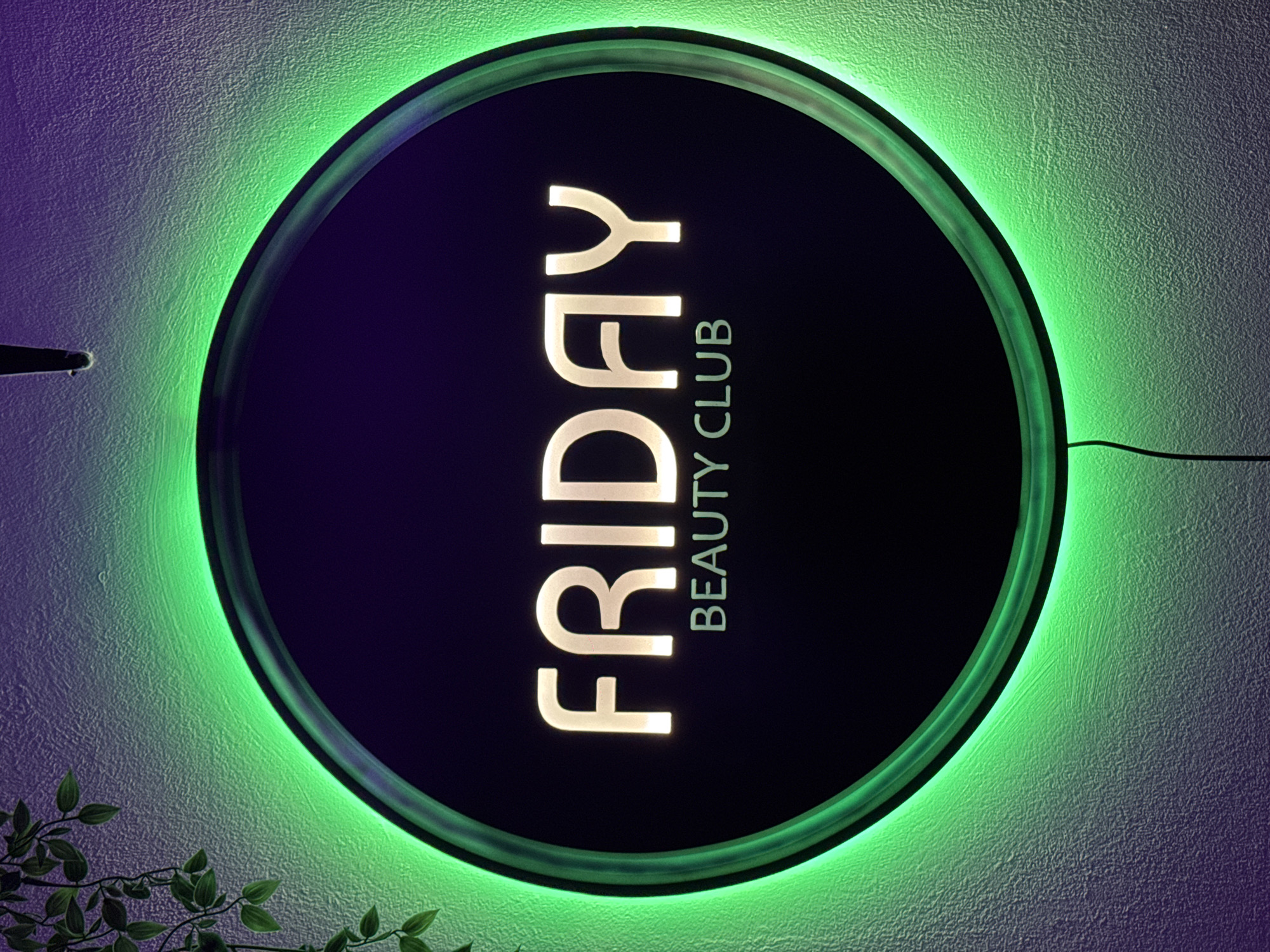 A circular, illuminated sign reading "FRIDAY BEAUTY CLUB" in vertical alignment hangs on a textured wall. The sign is backlit with green light, creating a halo effect around it. The interior of the circle is dark. A power cord is visible on the right side of the sign. A small plant with green leaves is visible in the lower-left corner of the image. The wall has a gradient of green to purple light. The location is likely Sofia, Bulgaria due to the name "FRIDAY BEAUTY CLUB" and its international connotations.