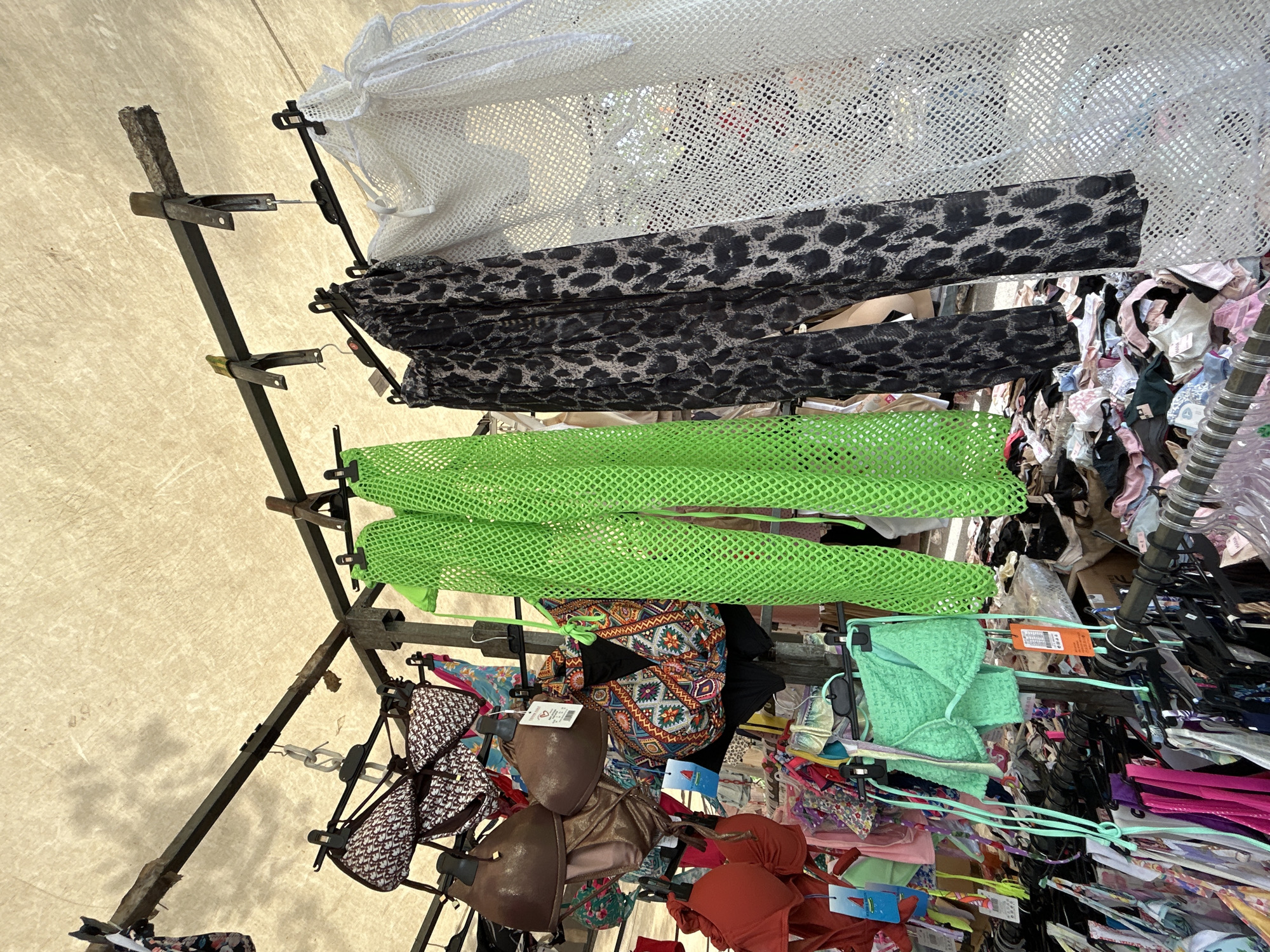 This is a medium shot, eye-level, outdoor photograph of a clothing stall, likely a market, under a beige awning. The stall displays a variety of swimwear and cover-ups hanging on metal racks. In the foreground, to the left, are several bikini tops and bottoms, some in solid red and green, others with patterned designs. Hanging centrally are a pair of lime green mesh pants and a matching bikini top, as well as a pair of black and white leopard print sheer pants. To the far right is a white lace-like sheer cover-up. The background shows more clothing items piled on the ground and hanging on racks, suggesting a busy retail environment. The overall scene is well-lit with natural light filtering through the awning.