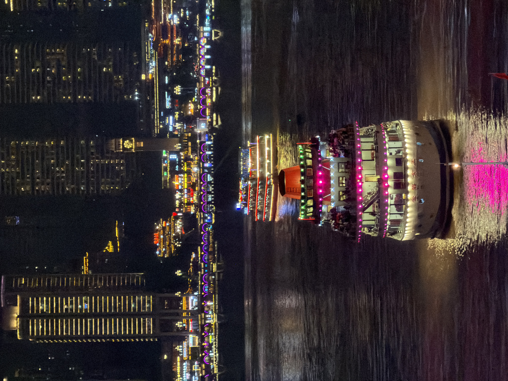 This is a nighttime image of a waterfront scene in Yuzhong District, China, likely taken from a boat or a high vantage point. The scene is dominated by the glow of city lights reflecting on dark water.
The primary focus is a brightly lit, multi-decked boat moving through the water. The boat is adorned with numerous colorful lights, predominantly pink and white, outlining its structure and windows. People are visible on the boat's decks, suggesting it is a tourist vessel or a party boat. The boat is creating a wake as it travels, with splashes of light from the boat illuminating the water around it.
To the left of the boat, the waterfront is lined with illuminated buildings and structures. These buildings display a variety of lights, ranging from warm yellow and white to vibrant purple and blue, creating a dazzling urban skyline. Some of these structures appear to be modern high-rise buildings with many windows, while others seem to be smaller, more ornate buildings or entertainment venues closer to the water's edge. The lights from these buildings are also reflected in the water, creating a shimmering effect.
The overall atmosphere is one of vibrant nighttime activity and urban entertainment. The time of day is clearly night, and the water appears calm, with ripples and reflections indicating movement. There is no indication of weather conditions. The location is identified as Yuzhong District, China, which is consistent with the dense urban development and waterfront activity depicted. There is no discernible text in the image.