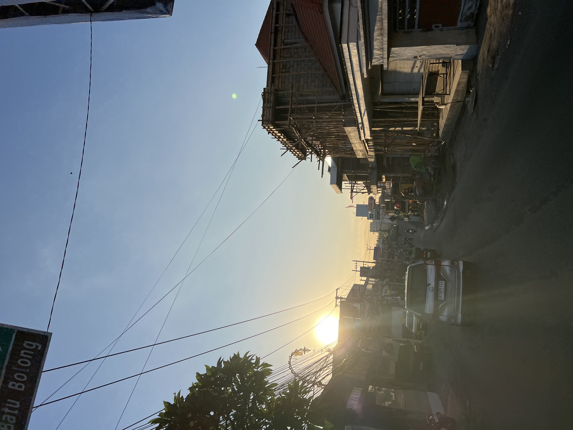 The image depicts a street scene in North Kuta, Indonesia, during what appears to be either sunrise or sunset, judging by the low angle of the sun and its bright, diffused light. The sky is a clear, pale blue with a slight gradient towards the horizon.
On the left side of the street, several buildings are visible, some of which appear to be shops or businesses. A sign overhead reads "Batu Bolong." A tree with dense green foliage is present on the far left. Several power lines crisscross the scene, contributing to a somewhat cluttered urban aesthetic.
The street itself is paved and appears to be a two-way road. Traffic includes a silver minivan in the foreground with a license plate that reads "DK 1679 FCS." Several motorcycles and other vehicles are also on the road, some of them moving.
On the right side of the street, a building is under construction. It features a wooden scaffolding structure surrounding a brick and concrete frame. Red-tiled roofs are visible on some of the finished or partially finished structures. Construction workers, dressed in casual clothing and one in a green vest, are present near the construction site.
The overall atmosphere is that of a bustling street in a developed area, with elements of both daily life and ongoing development. The sunlight creates strong highlights and shadows, contributing to a dynamic visual quality.