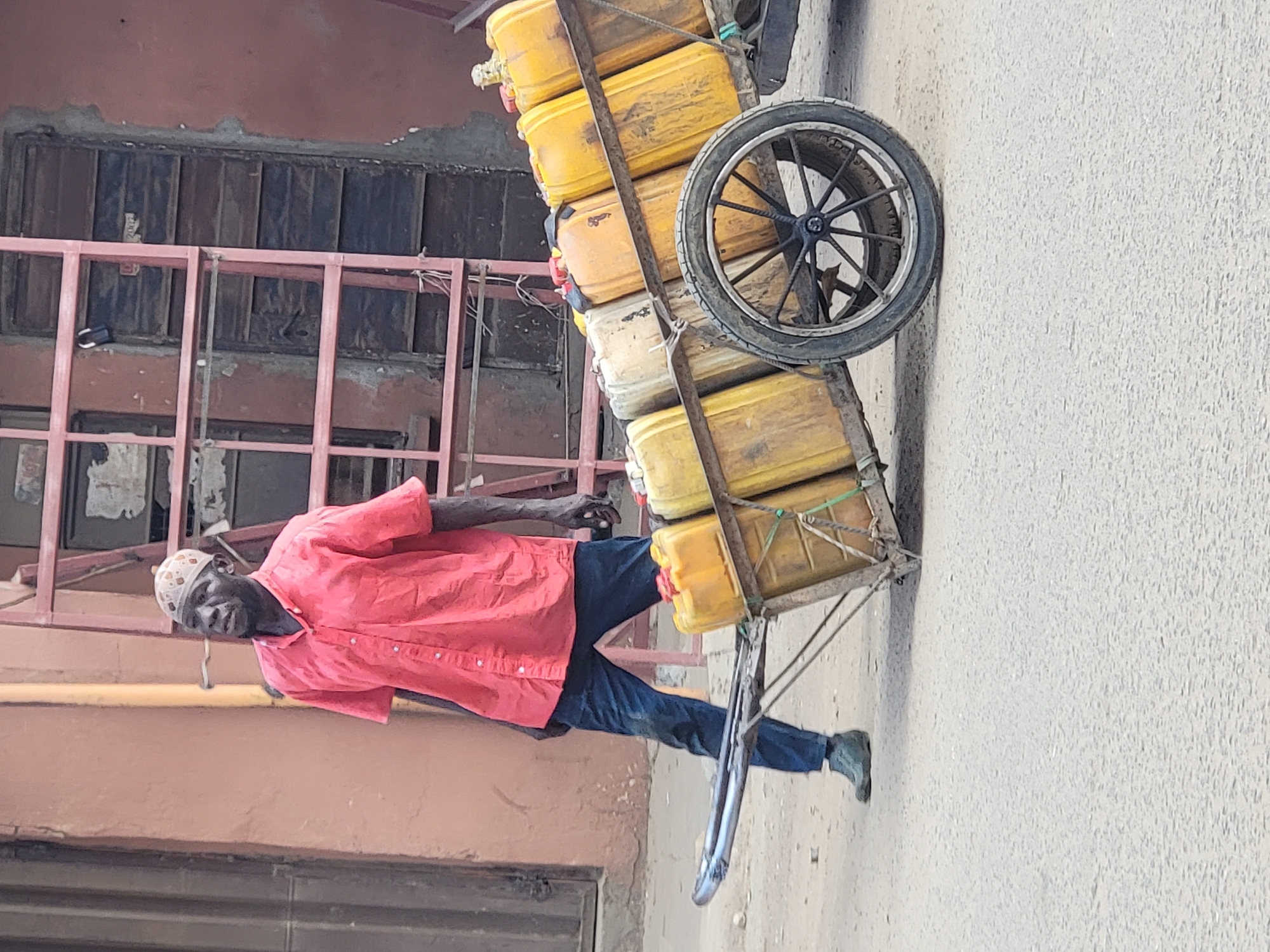 The image depicts a man in Maiburiji, Nigeria, pulling a cart loaded with yellow jerrycans of water. He is wearing a red button-down shirt, blue jeans, and a patterned skullcap. The cart has a single wheel and appears to be handmade. The background includes a building with a window and a pink metal structure. The man is pulling the cart on an asphalt road or path.