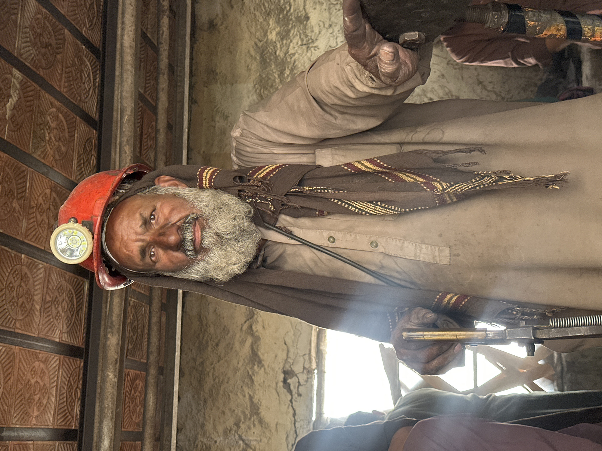 An elderly man with a grey beard, wearing a red hard hat with a headlamp, stands in what appears to be a mining or workshop environment in Quetta, Pakistan. He is wearing a light brown, button-down shirt and a dark brown scarf with yellow and red stripes. His right hand is gloved and rests on a piece of metal machinery. The background shows a rough, textured wall and a ceiling made of metal panels. The lighting suggests an indoor setting, possibly with natural light filtering in from the left. Other individuals are partially visible in the background, out of focus.