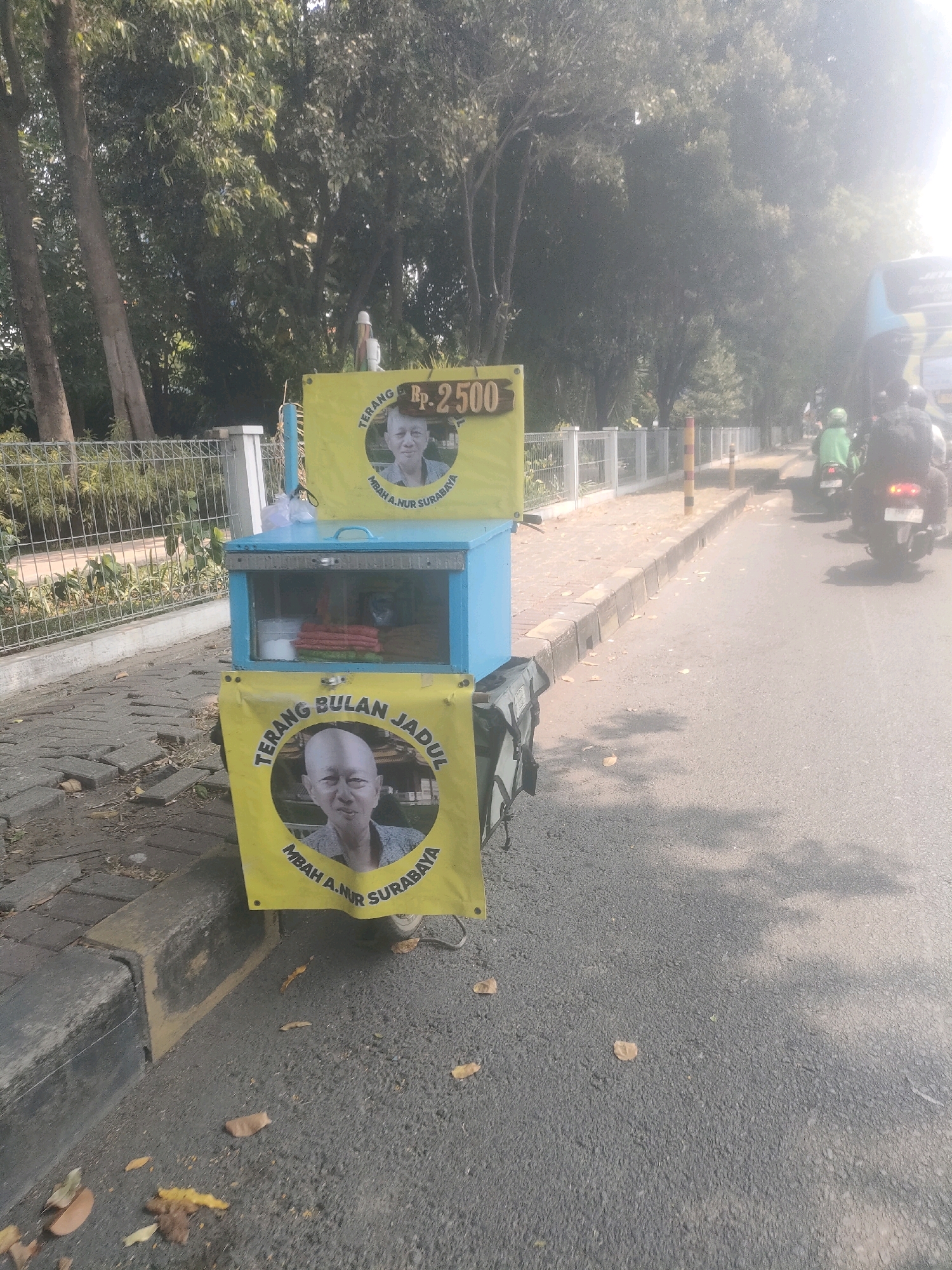This image shows a street vendor's cart on the side of a road in Surabaya, Indonesia. The cart is painted blue and has yellow signs with pictures of a bald man and the text "TERANG BULAN JADUL" and "MBAH A. NUR SURABAYA". The price "Rp. 2500" is also visible on one of the signs. The cart appears to be selling food, with containers and folded cloths visible inside the transparent section. In the background, there is a sidewalk with a metal fence, trees, and a road with traffic, including a bus and several motorcycles. The lighting suggests it is daytime, with sunlight casting shadows on the road.