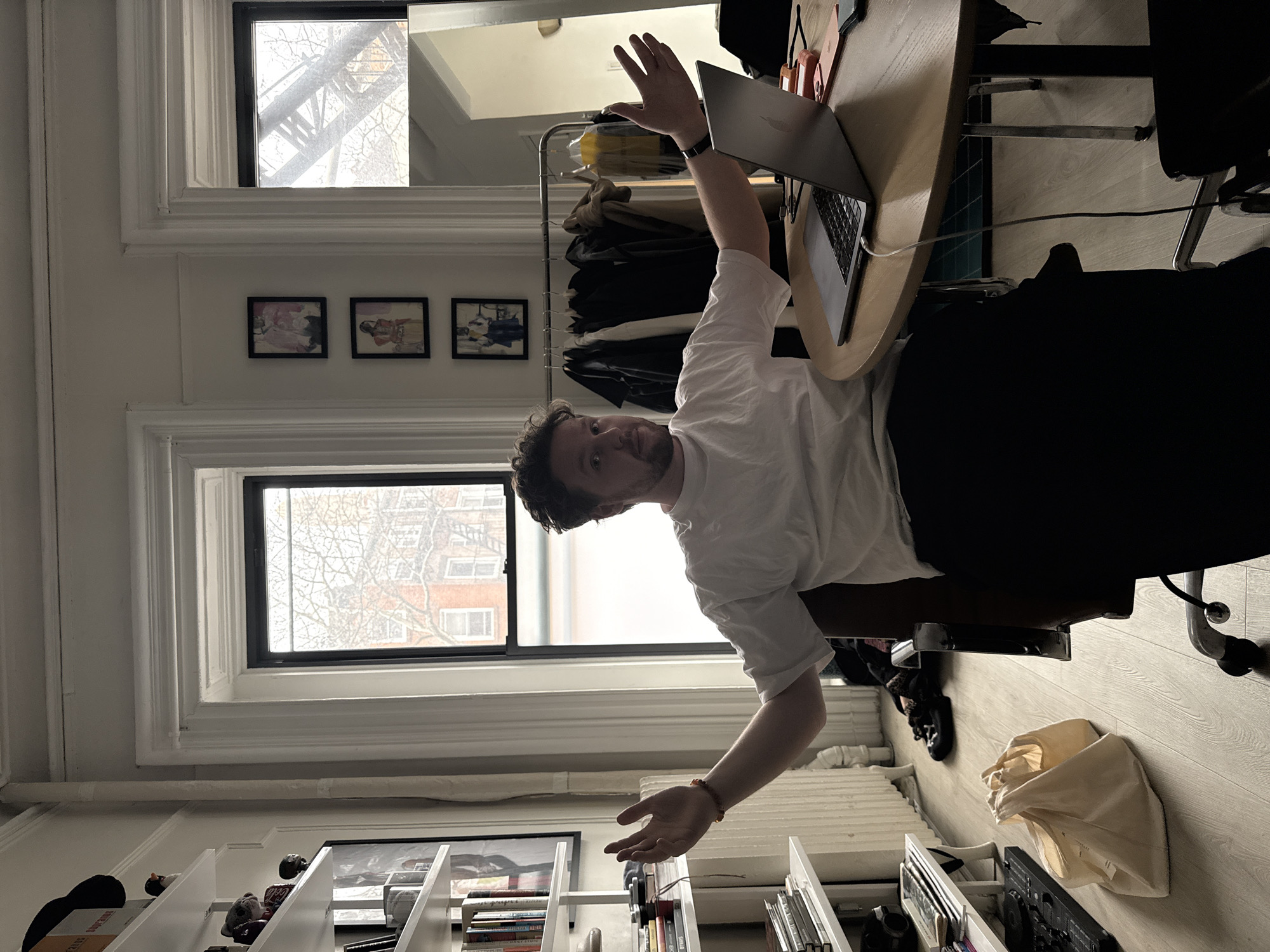 A man stands in what appears to be an apartment in New York City, facing the camera with arms outstretched. He is wearing a white t-shirt and dark pants. A laptop is on a round wooden desk to his right, with an orange phone and other items nearby. Behind him, clothes hang on a rack. Three framed pictures are mounted on the wall above the clothing rack. A window shows a view of brick buildings across the street. Below the window, on the right side of the frame, is a radiator. To the left, a white bookshelf is stocked with books, papers, and various objects. A white tote bag sits on the floor near a black chair. The room's flooring is light wood. Through the window, one can glimpse other apartment buildings, indicative of a densely populated urban area.