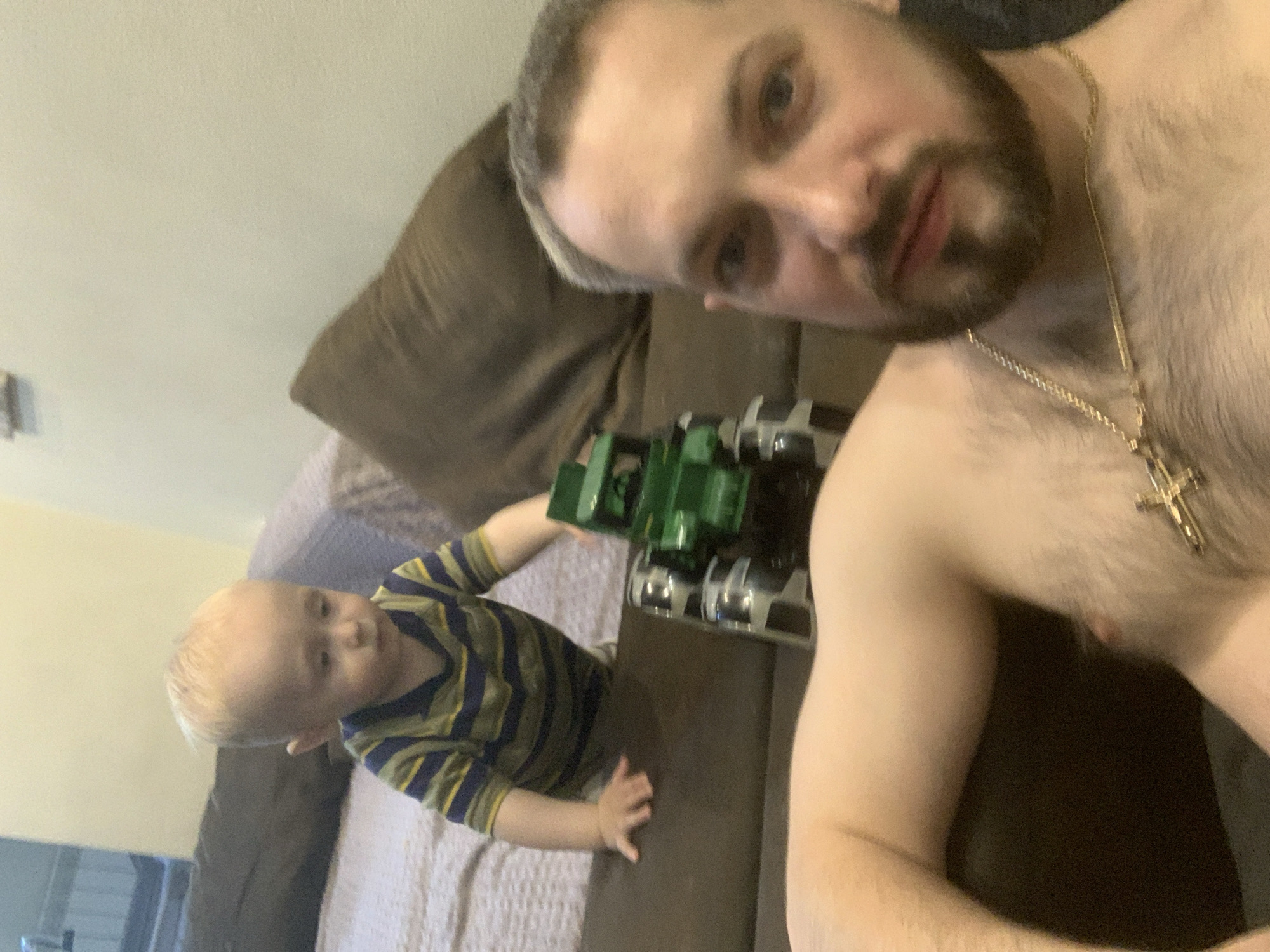 A man and a young child are on a brown sofa. The man is in the foreground, shirtless, with a gold chain and cross necklace. The child is in the midground, wearing a striped shirt, and is playing with a green toy tractor. The background consists of a wall and pillows.