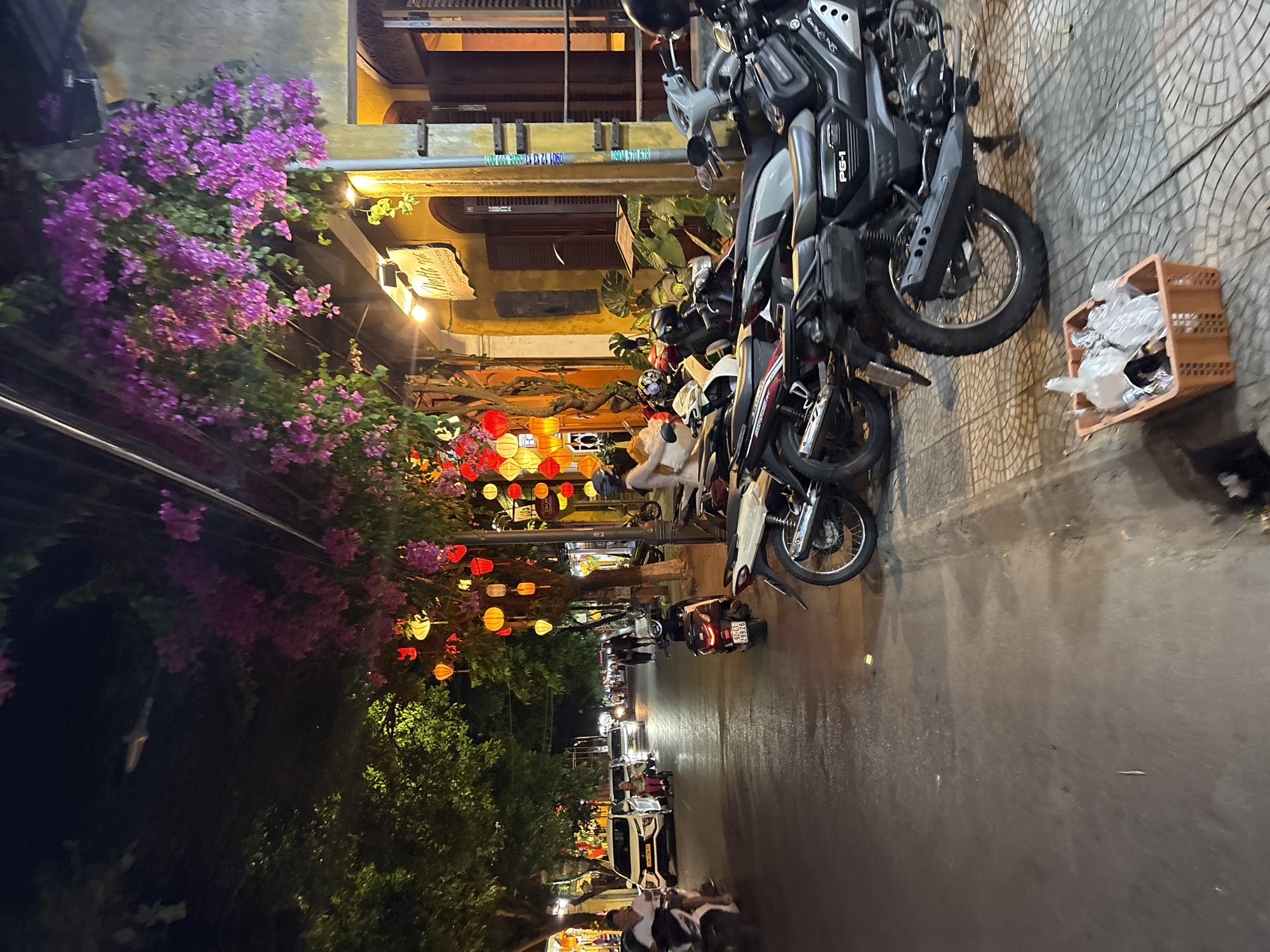 This photograph depicts a street in Hội An Ward, Vietnam, at night. The street is lined with parked motorcycles and scooters, some in close proximity. There are people walking along the street, visible in the distance and near the foreground. Buildings on one side of the street are adorned with colorful lanterns, and purple flowers hang from a structure. The street surface appears wet, possibly from recent rain. A small orange basket sits near a motorcycle on the paved sidewalk.