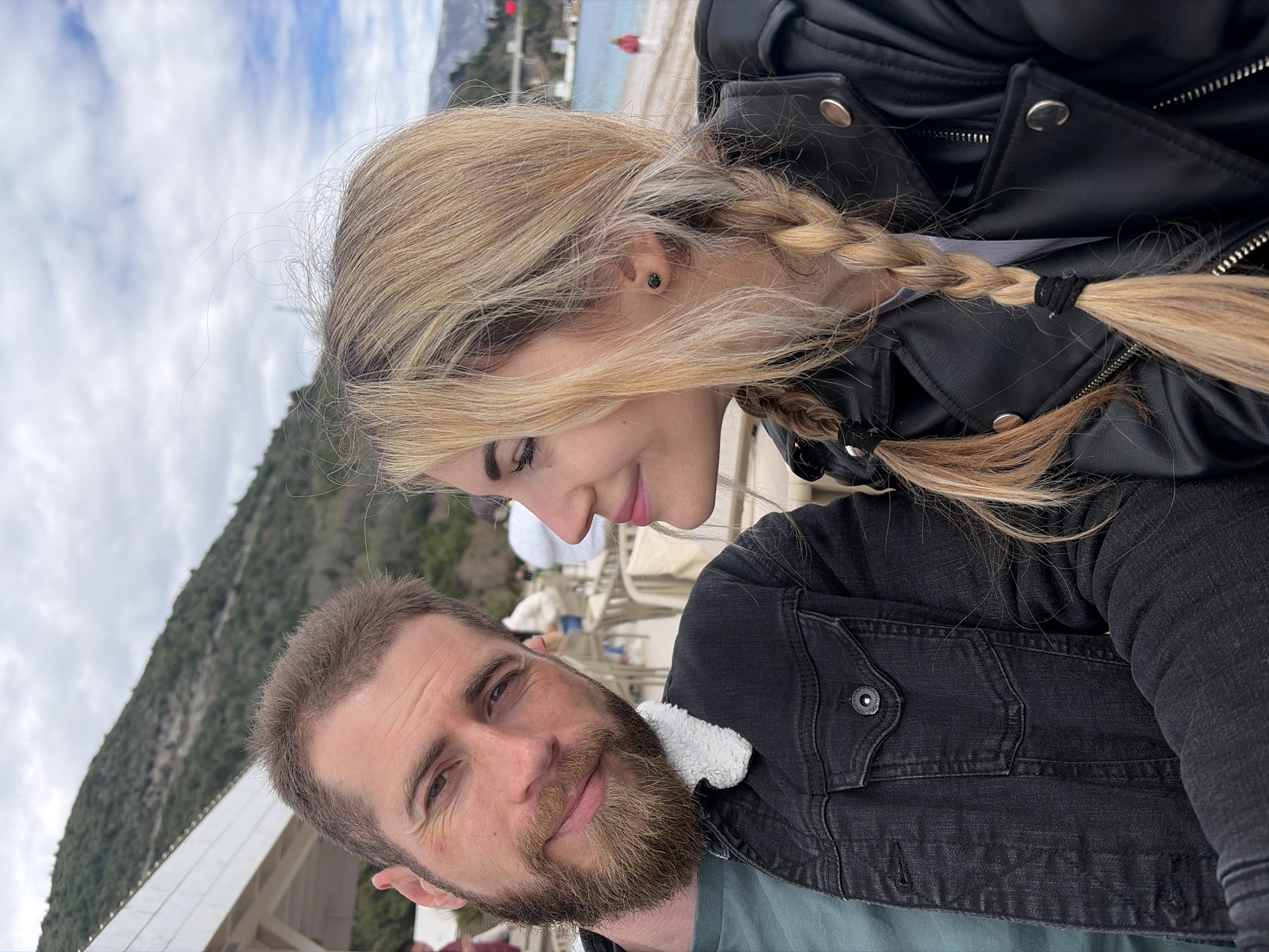 The image shows a couple in a selfie. The woman has blonde hair styled in braids, and is wearing a black leather jacket. She looks down towards the man. The man has a beard and is wearing a black denim jacket over a green t-shirt. Behind the couple is a mountain range, part of a white structure, and a glimpse of the sky with clouds. The location appears to be outdoors in Budva, Montenegro, possibly near the beach. Other individuals are faintly visible in the background.