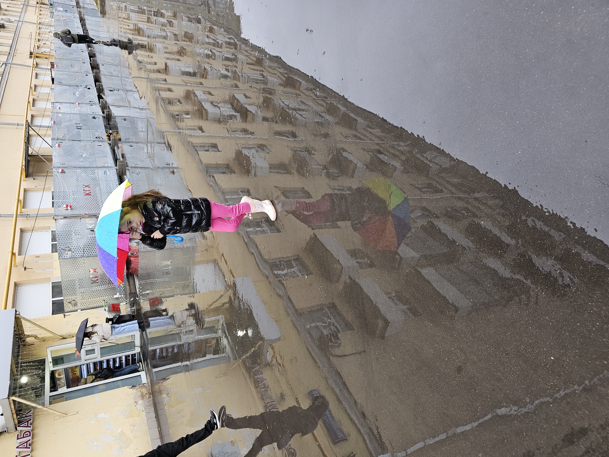 The image shows a reflection in a large puddle, likely after rainfall, in what appears to be an urban setting. The reflection features a multi-story building with scaffolding present on the facade, suggesting ongoing construction or renovation work.
A young girl holding a colorful umbrella is captured in the reflection; she is wearing a black jacket, pink leggings, and white boots. The reflected image also shows the legs of a person clad in black pants and sneakers entering the puddle's perimeter.
In the background, a shop with visible lettering "ТАБАК" (TOBACCO) suggests the presence of a convenience store. Another individual holding a dark umbrella stands near the shop's entrance.
Based on the metadata, the image was taken in Khimki, Russia.