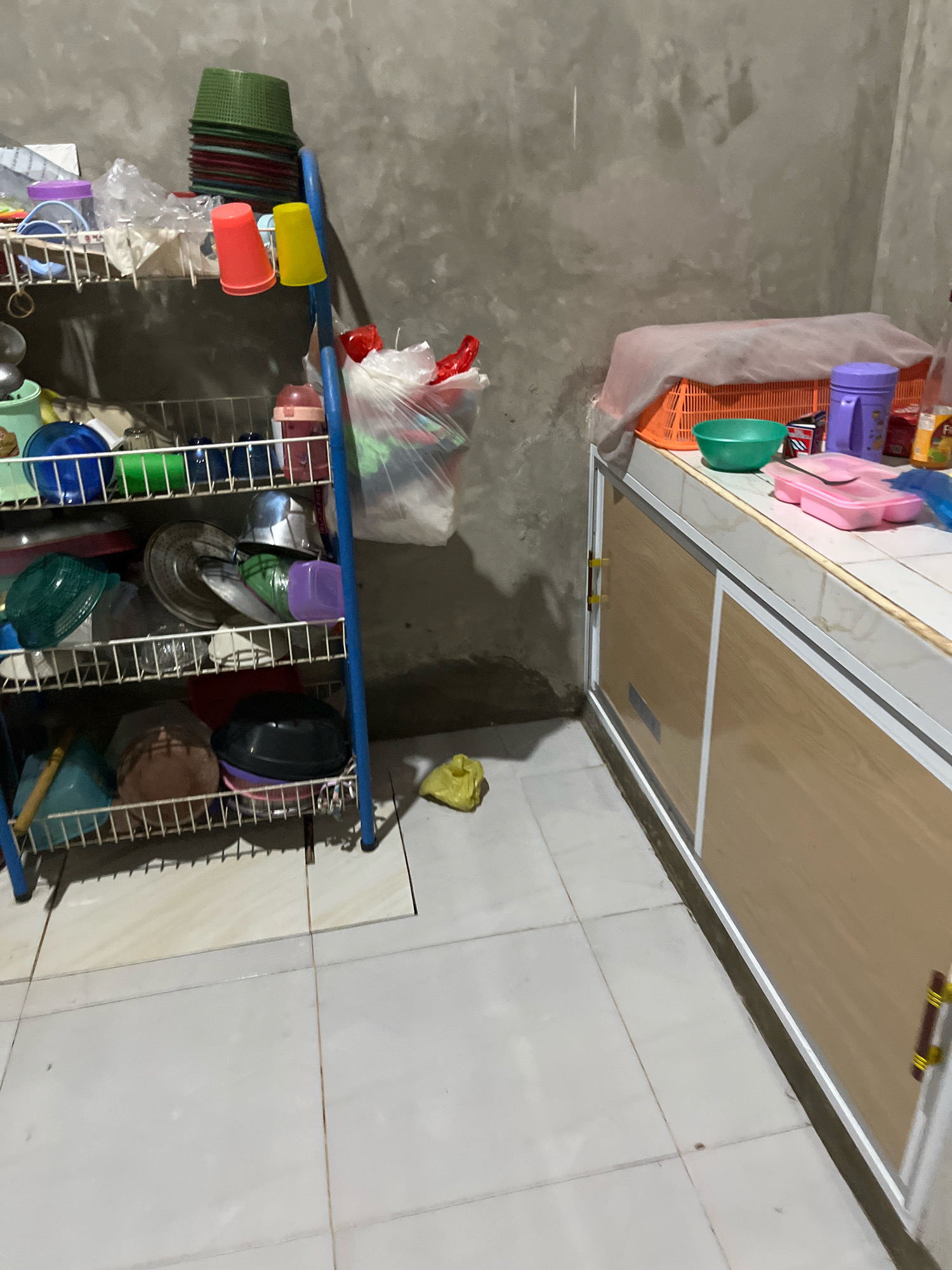 This is a medium-shot, indoor, eye-level, vertical, poorly lit photograph of a cluttered kitchen area in Kebumen, Indonesia. The scene shows a metal dish rack filled with various kitchen items and a cabinet with wooden doors on the right. The dish rack is blue and has multiple shelves holding plates, bowls, cups, and containers. Some of the items on the top shelf are red and green stacked bowls. On the shelf below, there are several plastic cups, two of which are red and yellow. A large plastic bag filled with groceries is hanging from the side of the dish rack. On the right side of the image, a kitchen cabinet with light-colored wooden doors and gold handles is visible. On top of the cabinet, there is an orange plastic basket covered with a pink cloth, a green bowl, and some purple containers. The walls are made of rough, gray concrete. The floor is tiled with light-colored square tiles. A crumpled yellow plastic bag is on the floor to the left of the cabinet. The overall impression is one of a working, functional kitchen that is somewhat disorganized.