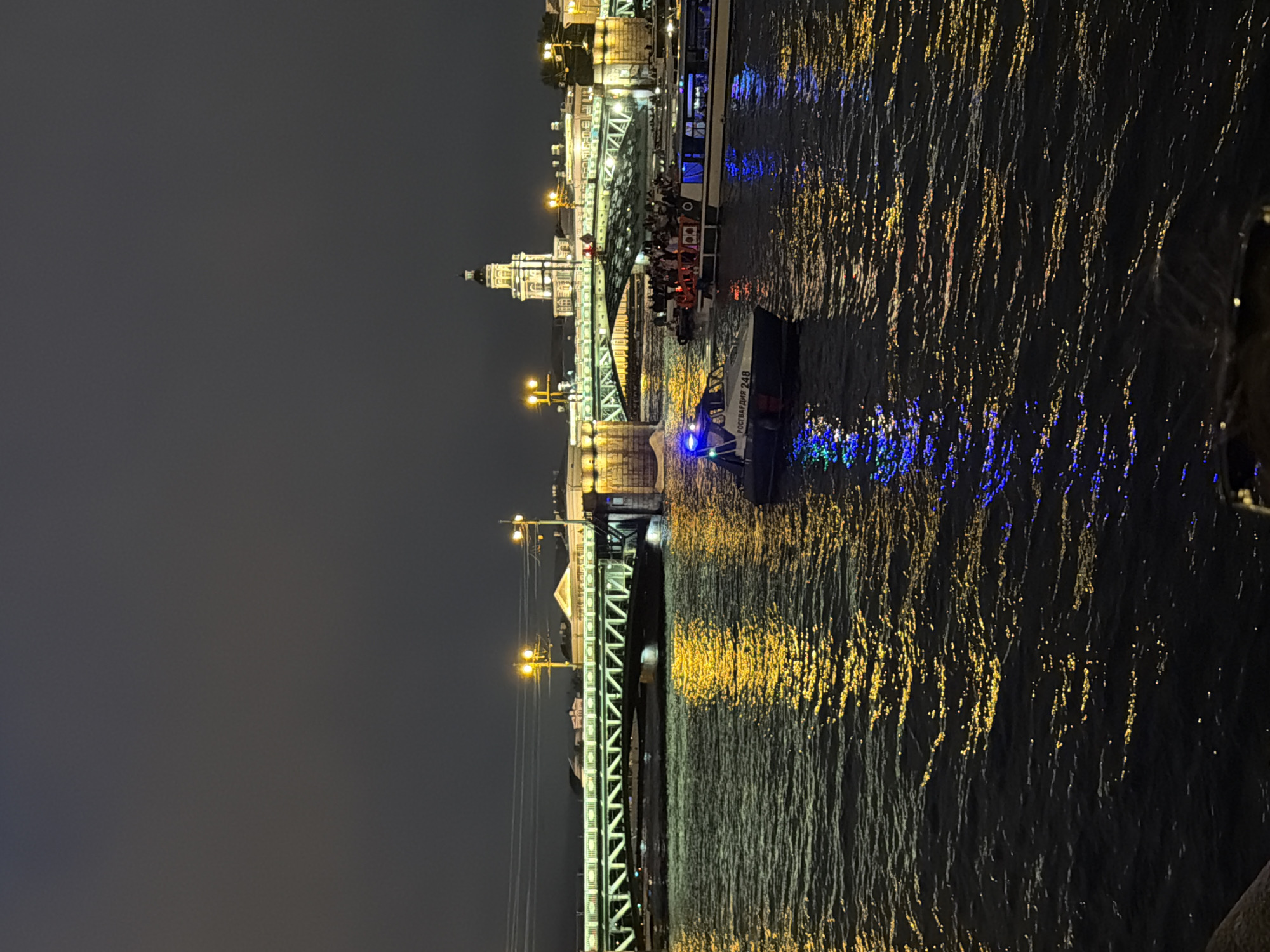 An evening scene captures a wide river with a bridge spanning across it, illuminated by lights. On the river, several boats are visible. In the foreground, a patrol boat with blue flashing lights is heading away from the viewer. Behind it, a larger boat filled with passengers, many wearing life vests, travels in the same direction. Further back, another boat, likely a tour boat, is also visible. The bridge is brightly lit, with yellow lights lining its structure and reflecting on the water. In the background, buildings and a tower with lights are silhouetted against the dark sky. The water's surface is rippled, with reflections of the bridge and boat lights creating a shimmering effect.