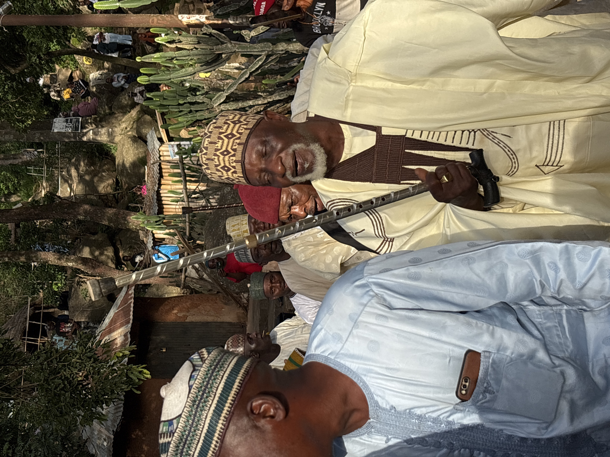 The image depicts a group of people gathered outdoors, possibly in a village or informal settlement in Jos, Nigeria. The scene appears to be set in a somewhat rustic environment with trees, rocks, and makeshift structures visible in the background. Some people are seated on the ground, while others are standing.
The focal point of the image is an elderly man wearing a traditional cap and a cream-colored robe with brown embellishments. He is holding a long, slender object that resembles a cane or staff. His eyes are closed, and he appears to be relaxed or possibly sleeping. Another man, wearing a red cap, is partially visible behind him, also with his eyes closed. In the foreground, another man is seen from the back, wearing a light blue robe and a patterned cap. A mobile phone with a brown case is tucked into his robe pocket.
The presence of what appears to be a traditional ceremony or gathering is suggested by the attire and the communal setting. The lighting indicates it might be daytime, with dappled sunlight filtering through the trees. The overall atmosphere seems peaceful and relaxed, with individuals seemingly engaged in quiet contemplation or rest. Some signs with text are visible in the background, but they are not clear enough to be deciphered. The environment suggests a warm climate.