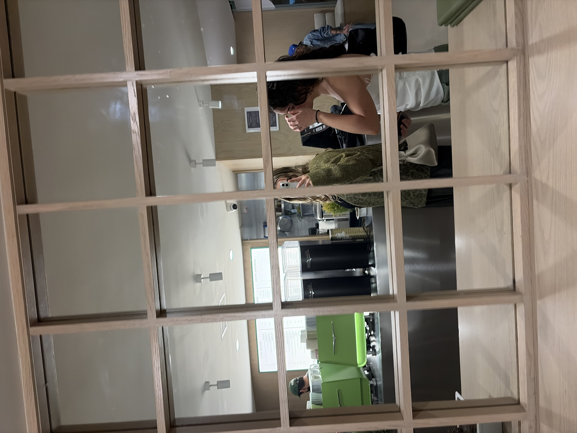 The image shows a reflection in a window with a light wood frame. The reflection shows the interior of a cafe or restaurant. Two women are visible; one is seated and appears to be looking down, possibly at her hands. The other woman is standing, holding a phone, and taking a picture. There's also a partial view of someone sitting in a chair near the seated woman. The interior includes menu boards, coffee machines, and green chairs. It's impossible to confirm the location in Puebla, Mexico, but the style of the interior and the people suggest a modern cafe or restaurant.