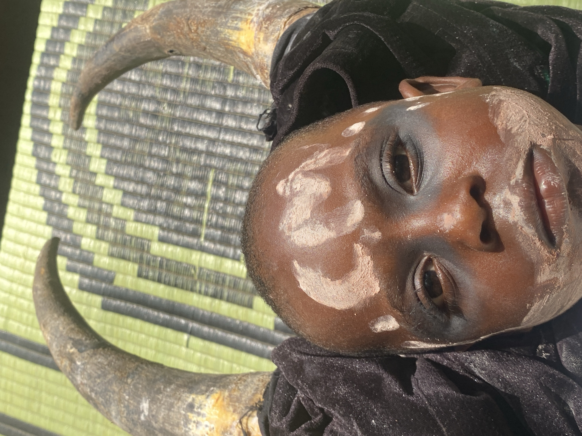 A child, painted with white markings, lies on a woven mat alongside two animal horns. The child is wrapped in a black cloth, obscuring the body. The scene is in Ufu Esioni, Nigeria.