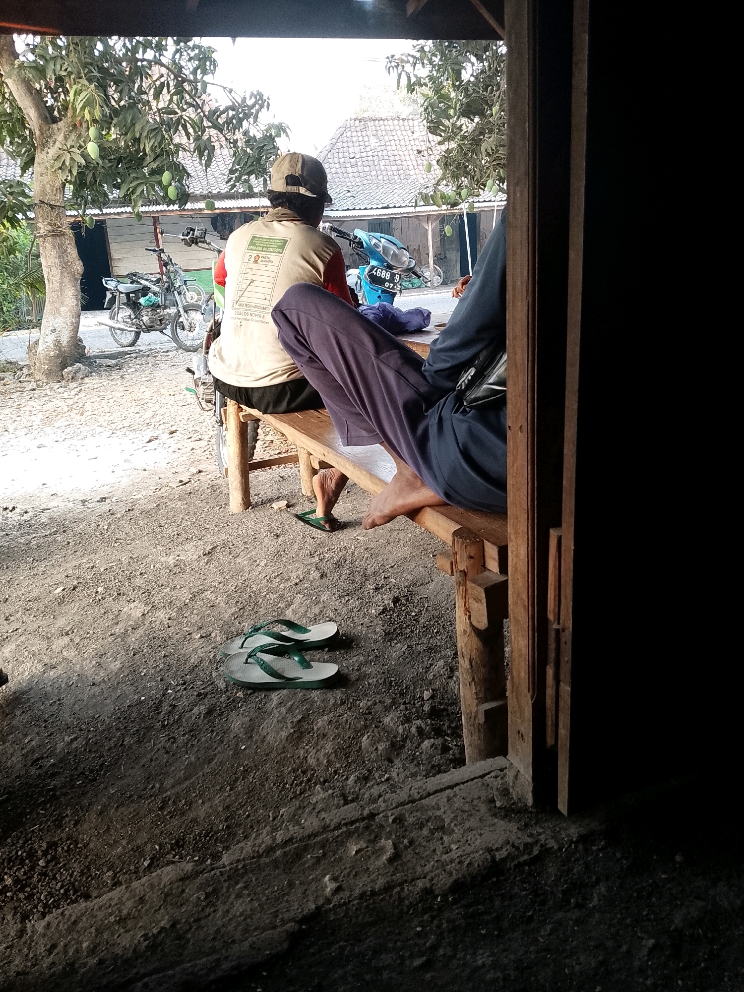 The image shows a rural scene in Wadang, Indonesia. In the foreground, a pair of light-colored flip-flops with green straps lies on the dirt ground. Behind them, a wooden bench is set up outdoors, with two individuals seated on it. The person on the left is seen from behind, wearing a light-colored long-sleeved shirt with text and graphics on the back, dark shorts, and a baseball cap. They are sitting on the bench and appear to be looking towards a motorcycle parked nearby. The motorcycle is partially visible and has a blue fuel tank. To the right of this individual, another person is seated on the bench, wearing dark purple pants and a dark blue long-sleeved shirt. Their legs are crossed, and their feet are visible, with one foot wearing a green flip-flop. A portion of a motorcycle is also visible behind the second individual, and a dark blue bag is resting on the bench.
The background features a dirt road with gravel and some vegetation. A tree with green mangoes is prominent on the left side of the image. In the distance, houses with tiled roofs are visible. The overall environment suggests a simple, outdoor setting in a village. The lighting indicates it is daytime, with sunlight casting shadows. The image is framed by a dark, wooden structure on the right, possibly part of a doorway or a porch.