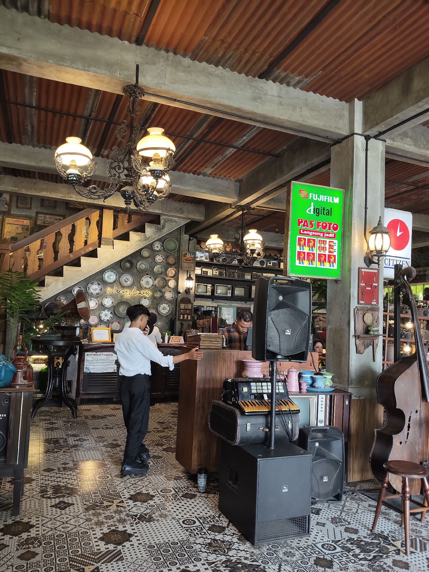 This image depicts the interior of a cafe or establishment in the City of Medan, Indonesia, characterized by its vintage and rustic decor. A young man in a white shirt and black trousers stands near a wooden counter, his right arm extended towards it. Behind him, a decorative staircase with wooden railings ascends. The walls are adorned with a collection of plates and framed pictures, contributing to the antique ambiance. Above, industrial-style corrugated metal roofing is visible, supported by concrete beams. Several ornate chandeliers hang from the ceiling, casting a warm light.
In the foreground, large black speakers and a keyboard are set up, suggesting a performance or entertainment area. A double bass and a wooden stool are also present, further enhancing the establishment's unique character. A prominent green sign with "FUJIFILM digital PAS FOTO LANGSUNG JADI!" advertising photo services is mounted on a pillar. The floor is covered with an intricate patterned tile, adding to the overall aesthetic. The scene captures a moment of stillness, with patrons potentially out of frame or subtly present in the background.