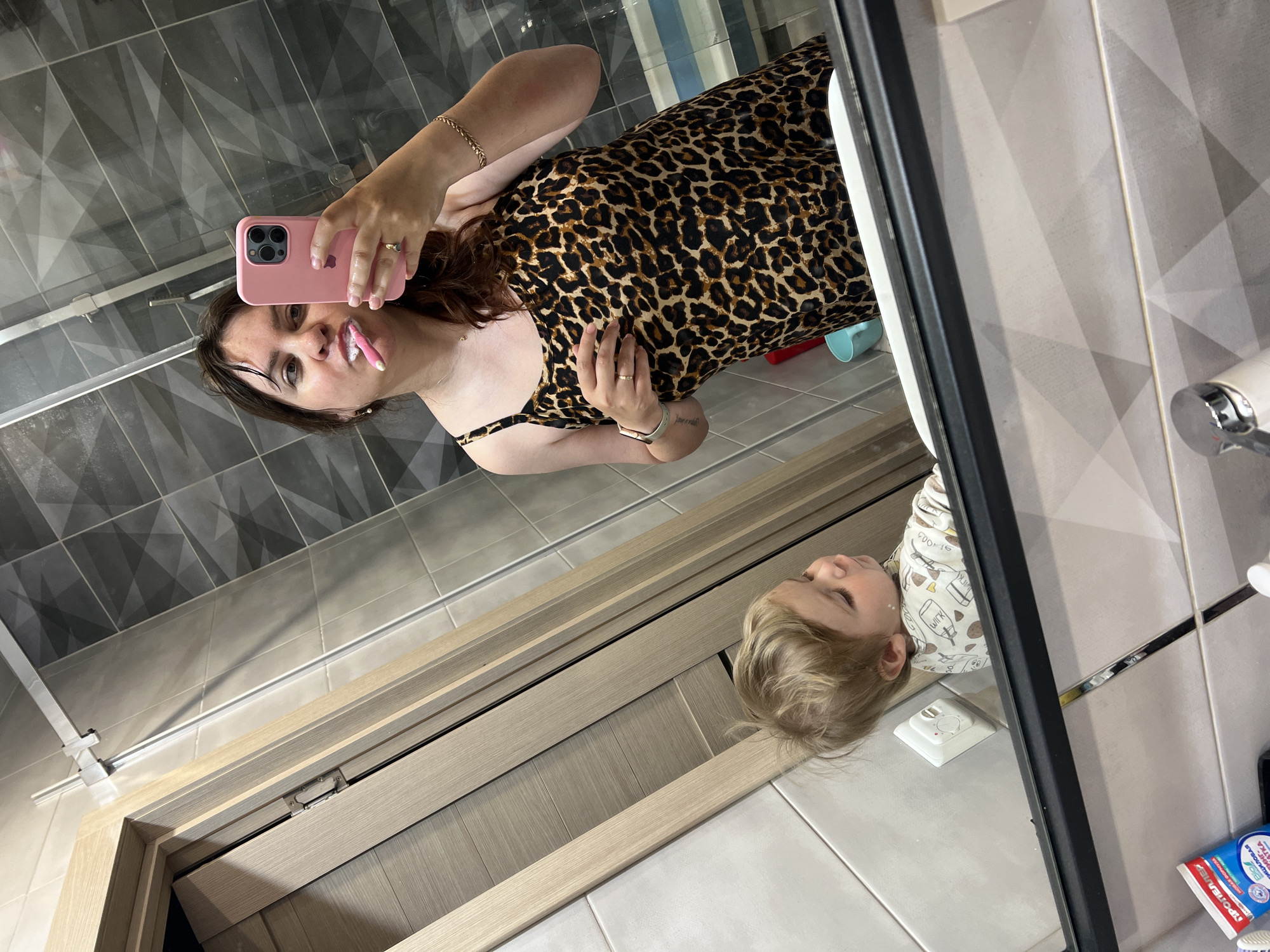 In a bathroom setting, a woman with dark hair, wearing a leopard print dress, is reflected in a mirror as she brushes her teeth with a pink toothbrush. She holds a pink smartphone in her right hand, also reflected. Beside her in the reflection, a young child with blonde hair is visible, looking downwards. The bathroom features grey tiled walls with a geometric pattern and a wooden door or cabinet. The overall scene appears to be a candid moment captured in a residential bathroom in Noviy, Russia.