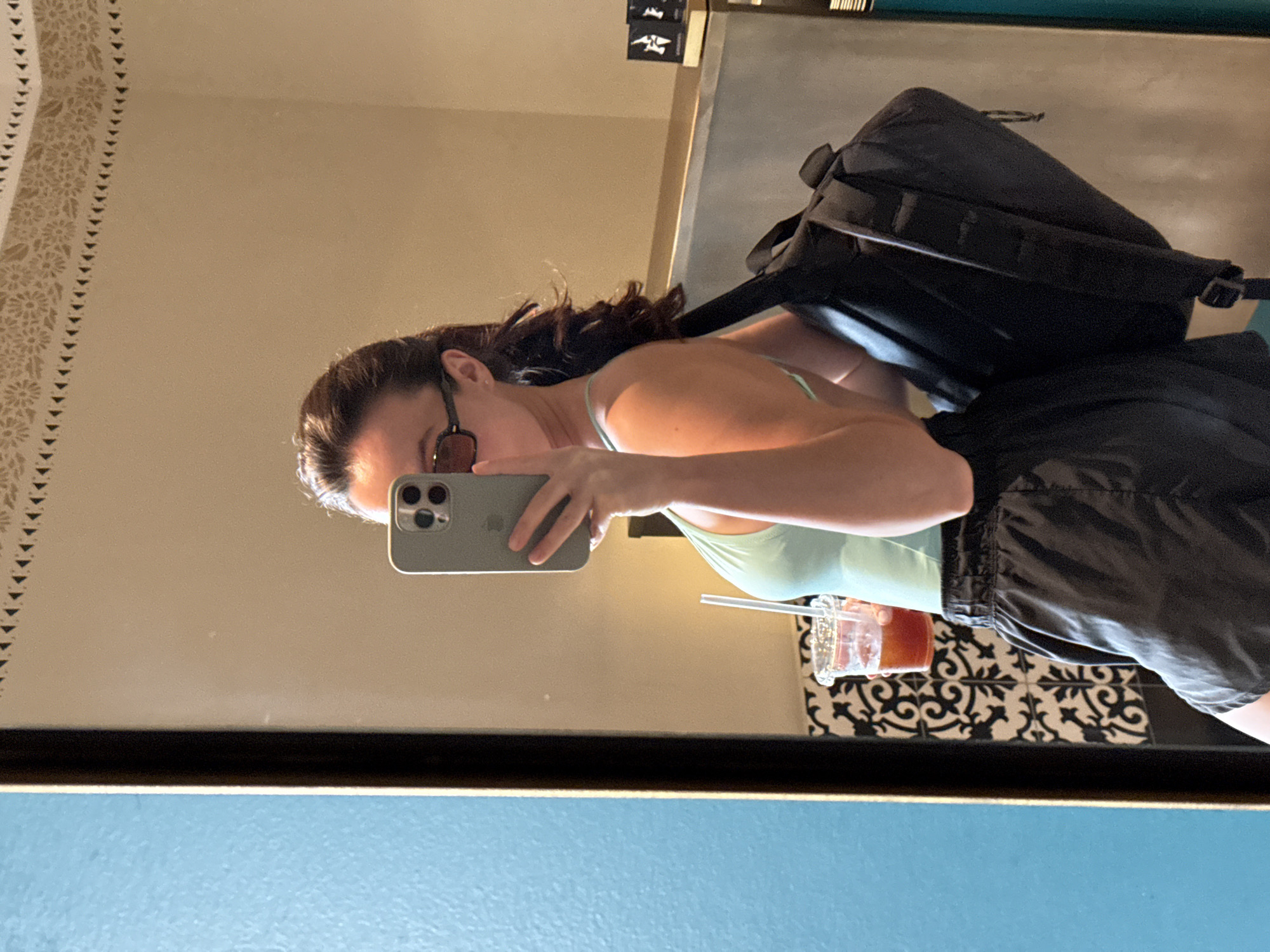 A young woman with dark hair, wearing sunglasses, a light blue tank top, and black shorts, is taking a mirror selfie. She is holding a clear plastic cup with a straw and a reddish beverage in her left hand and a black backpack slung over her right shoulder. The background appears to be a hotel room in Tel Aviv, Israel, with patterned tiles on the wall and a decorative border along the top. The woman is positioned in front of a mirror, which reflects her and the surrounding environment.