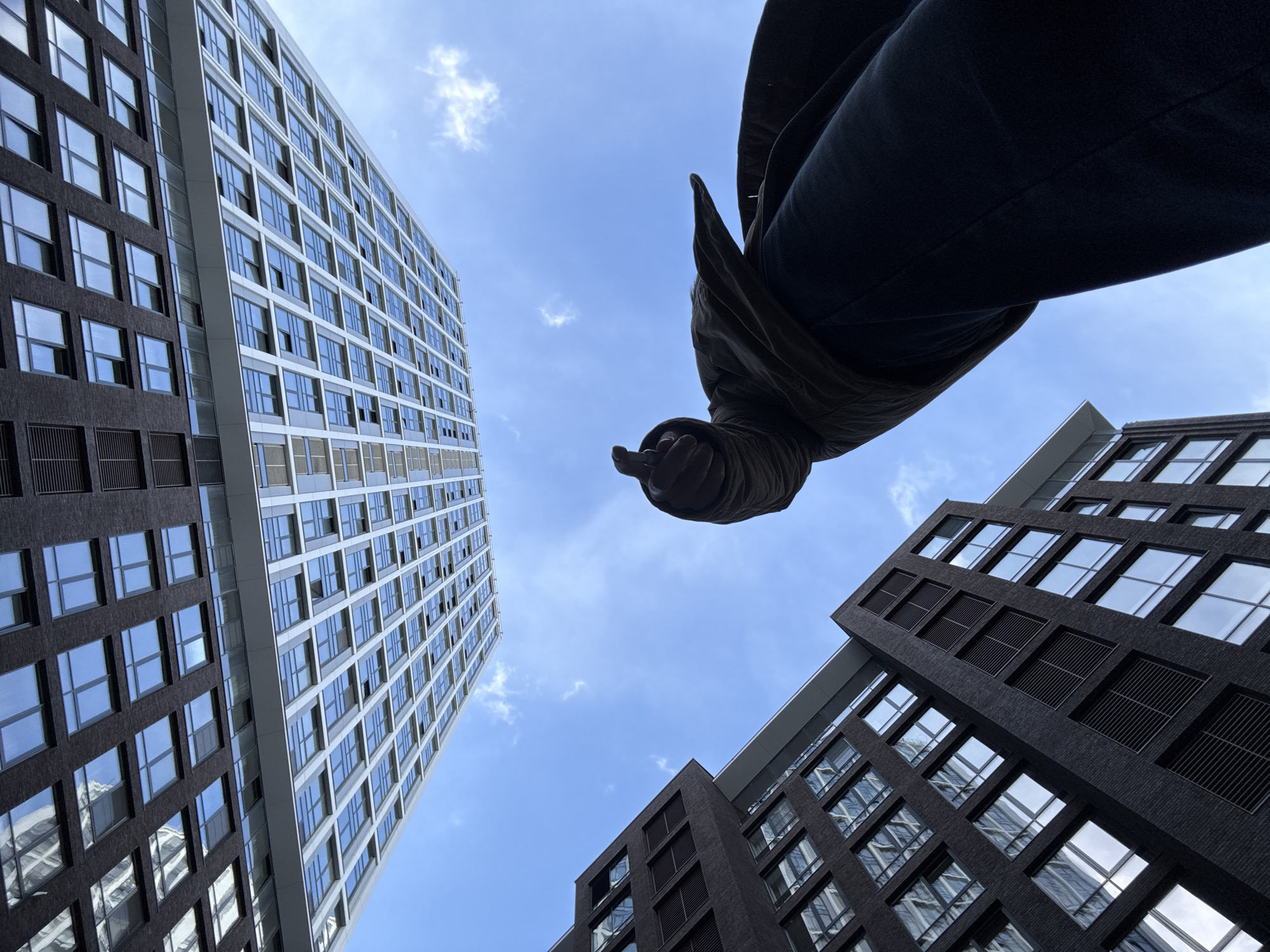 The low-angle shot features two buildings rising toward a partly cloudy blue sky. One building is light-colored with numerous windows in a grid pattern. The other building is darker, with a similar grid of windows. A person's lower body, clad in dark clothing and a jacket, partially obscures the sky. The person's hand is visible, reaching downward. The image was taken in Moscow, Russia.
