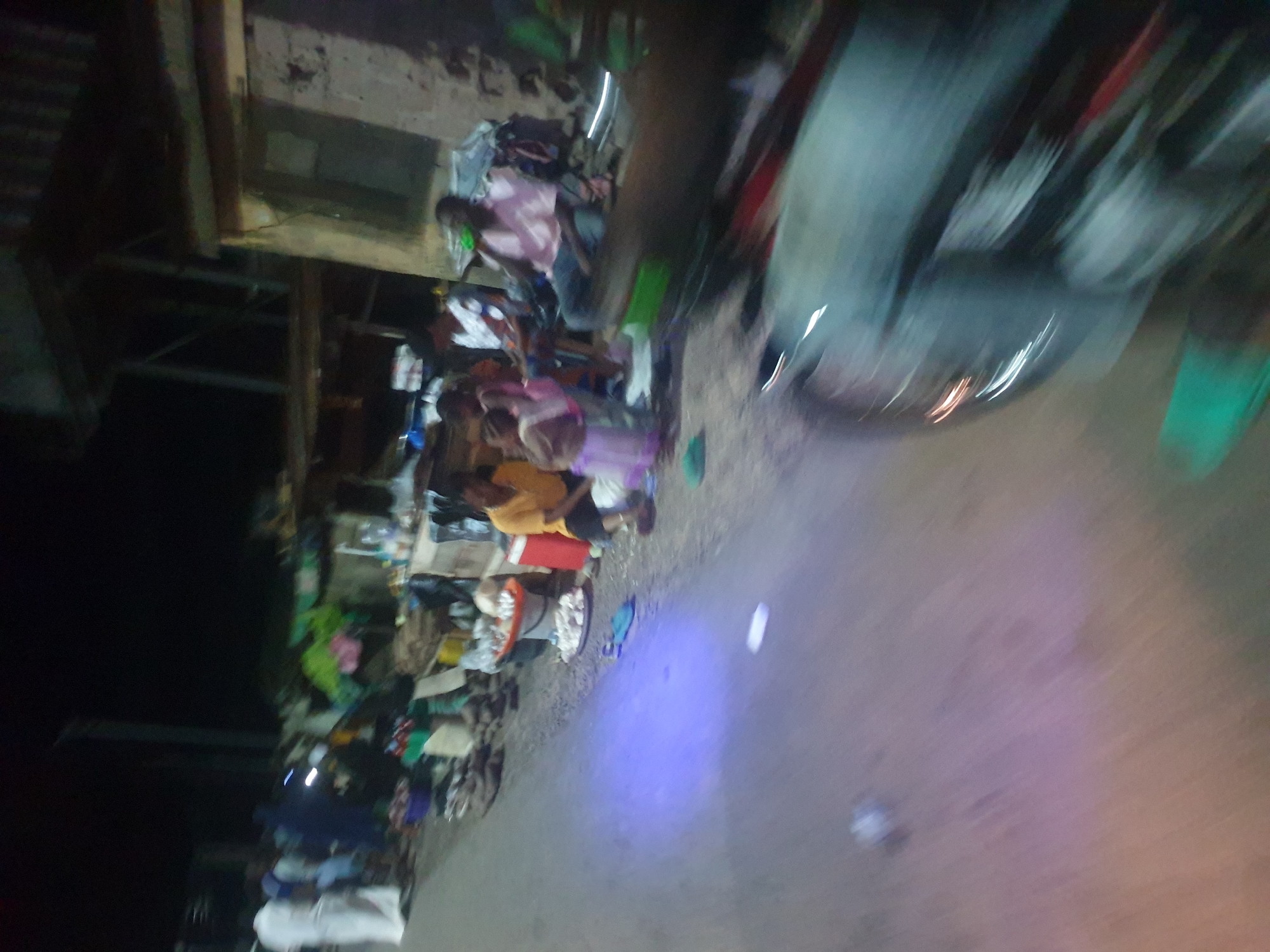 The image depicts a nighttime street scene in Zango-ndaji, Nigeria. Along the roadside, there are several people, possibly vendors, with goods displayed on tables and the ground. The objects include clothing, food items, and other wares. There is a vehicle parked on the road, partially obscuring the view. The environment is dimly lit, suggesting limited street lighting. The setting appears to be a bustling commercial area along a street.