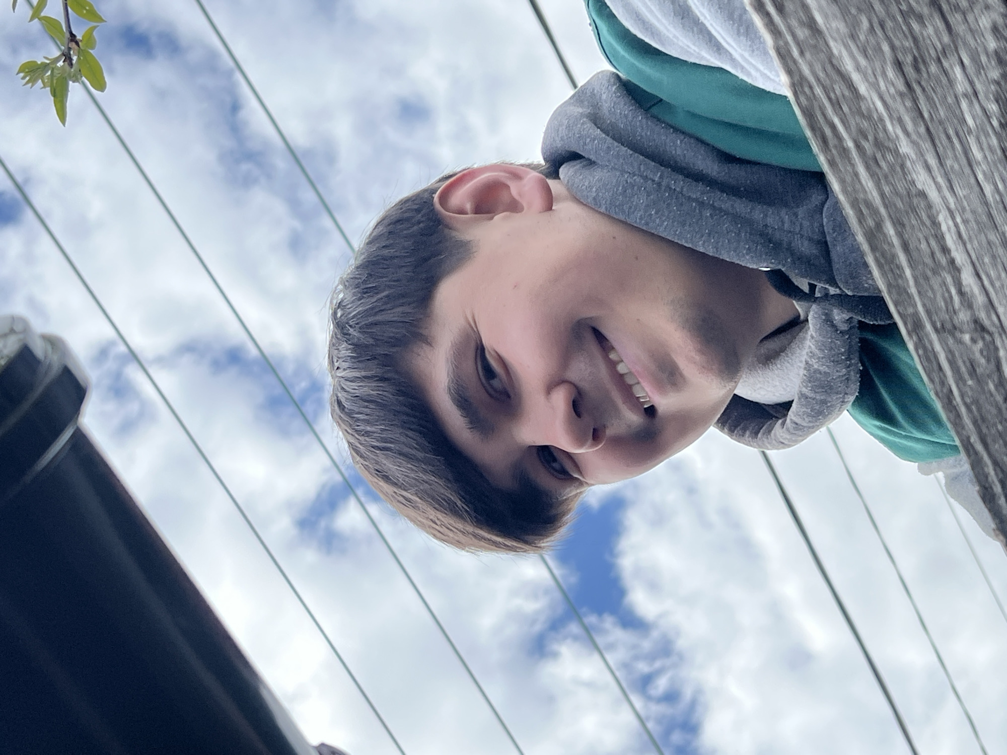 A low-angle close-up photograph depicts a smiling young man with short dark hair, wearing a grey and green hooded sweatshirt. The background shows a cloudy sky with patches of blue. In the foreground, there is a weathered wooden structure and dark-colored metallic or plastic object. The location in the city of Chaikovsky, Russia is not discernible from the visual information provided.